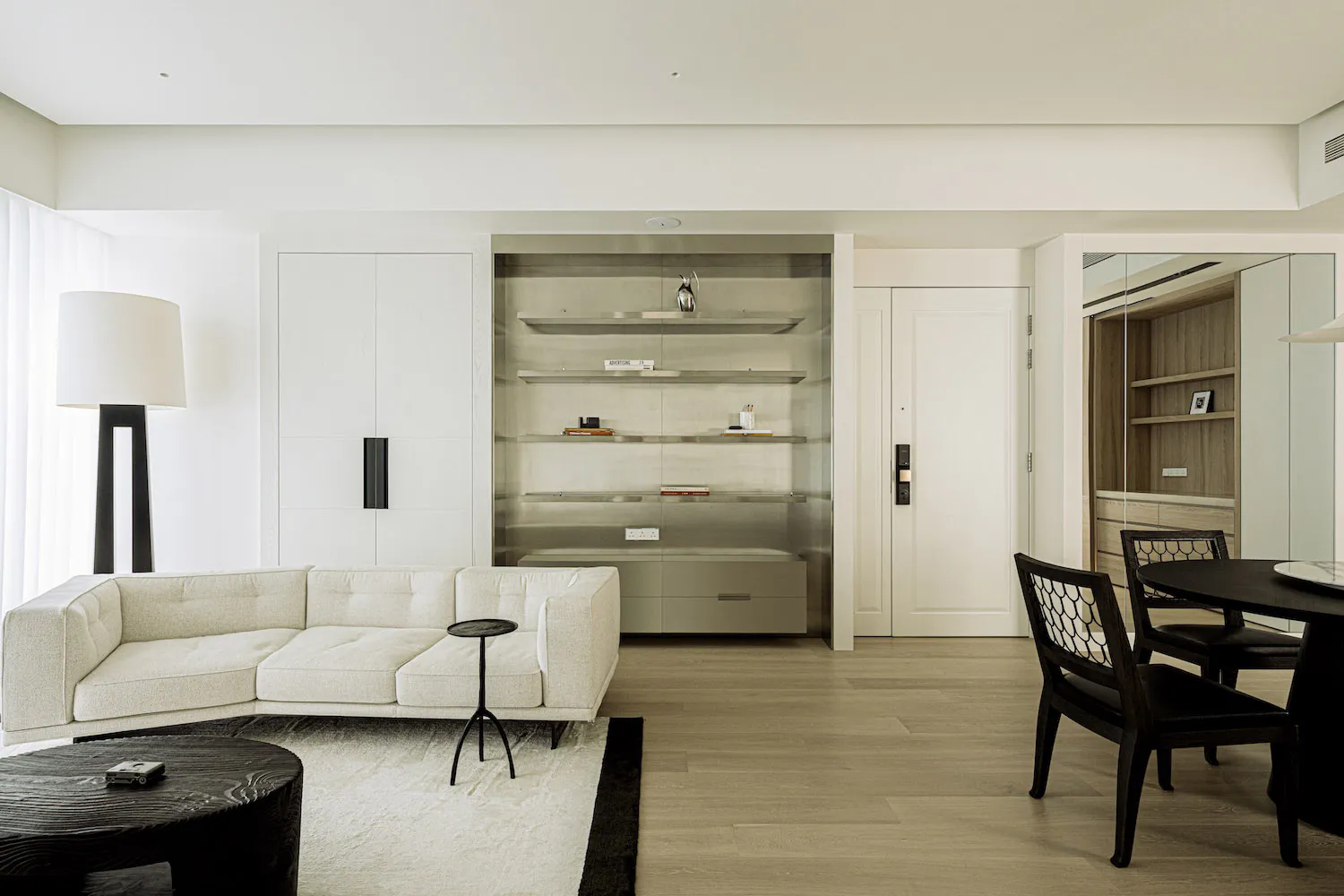 Modern minimalist living room with white L-shaped sectional sofa, dark round coffee table on white rug, built-in glass shelving unit with white oak cabinetry, and dark dining chairs around a round table.