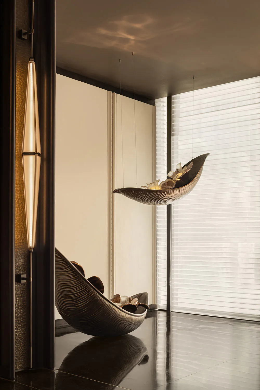 Modern luxury interior featuring a sculptural bronze leaf-shaped hanging lounge chair suspended from the ceiling and a matching floor version, with a tall pendant light on the left and large windows with sheer blinds in the background.