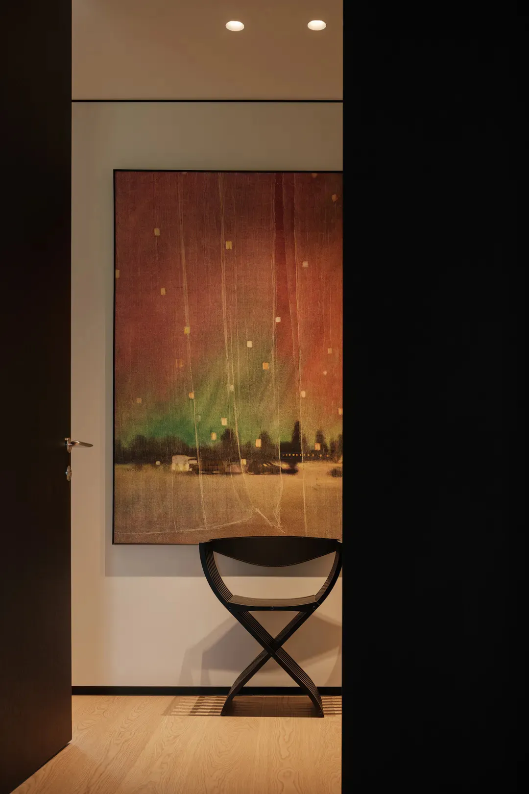 Modern entryway featuring a large vertical abstract painting with red, green, and gold tones depicting a landscape with hanging lanterns, paired with a black X-shaped stool on light wood flooring.