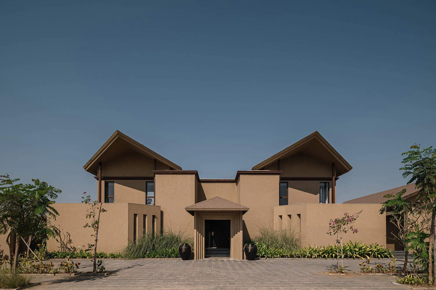 Front exterior view of a modern minimalist villa with beige sandstone walls, symmetrical triangular gables, central entrance portico, and desert-style landscaping with ornamental grasses and flowering trees.