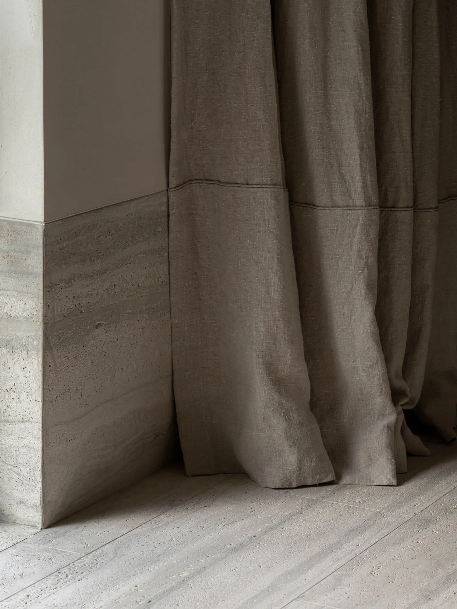 Close-up of a heavy natural linen curtain in muted taupe hanging beside a two-tone wall of smooth plaster above and layered travertine stone below, resting on a matching travertine floor.