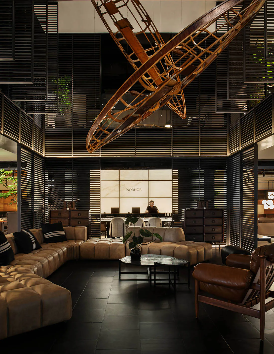 Modern luxury hotel or office lobby with dark wood slat walls, a large curved brown leather sectional sofa, matching leather armchair, round coffee tables, and a striking large-scale wooden biplane sculpture suspended from the ceiling. A reception desk with the brand name NORHOR is visible in the background.