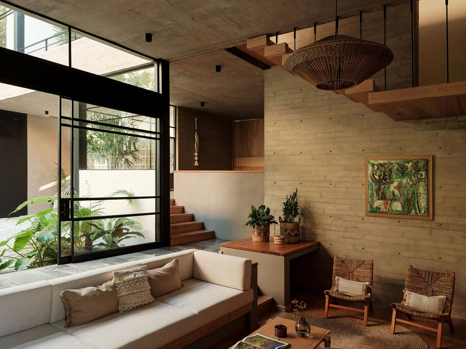 Modern living room featuring exposed concrete walls, a large woven rattan pendant lamp, two rattan lounge chairs, a built-in concrete sofa with beige cushions, wooden accents, and lush greenery visible through large black-framed glass doors.