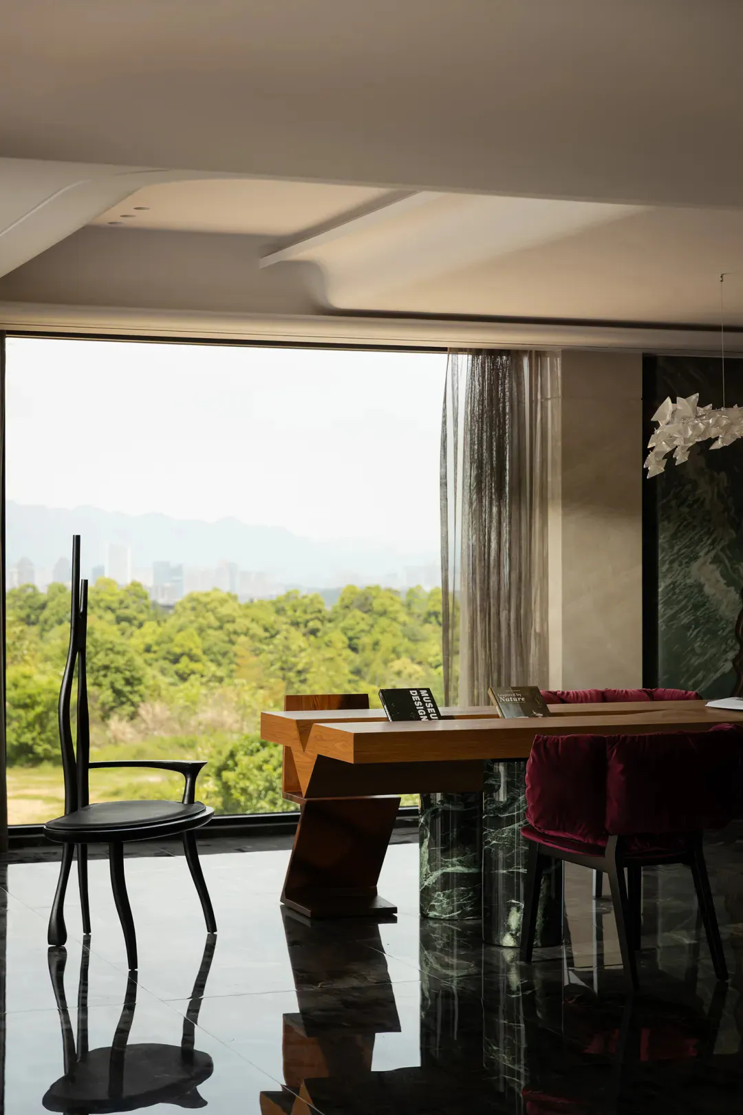 Modern dining room featuring a sculptural wooden dining table with a green marble cylindrical base, black organic-shaped chair, and burgundy velvet dining chairs, set against a floor-to-ceiling window overlooking a lush green landscape and distant city skyline.