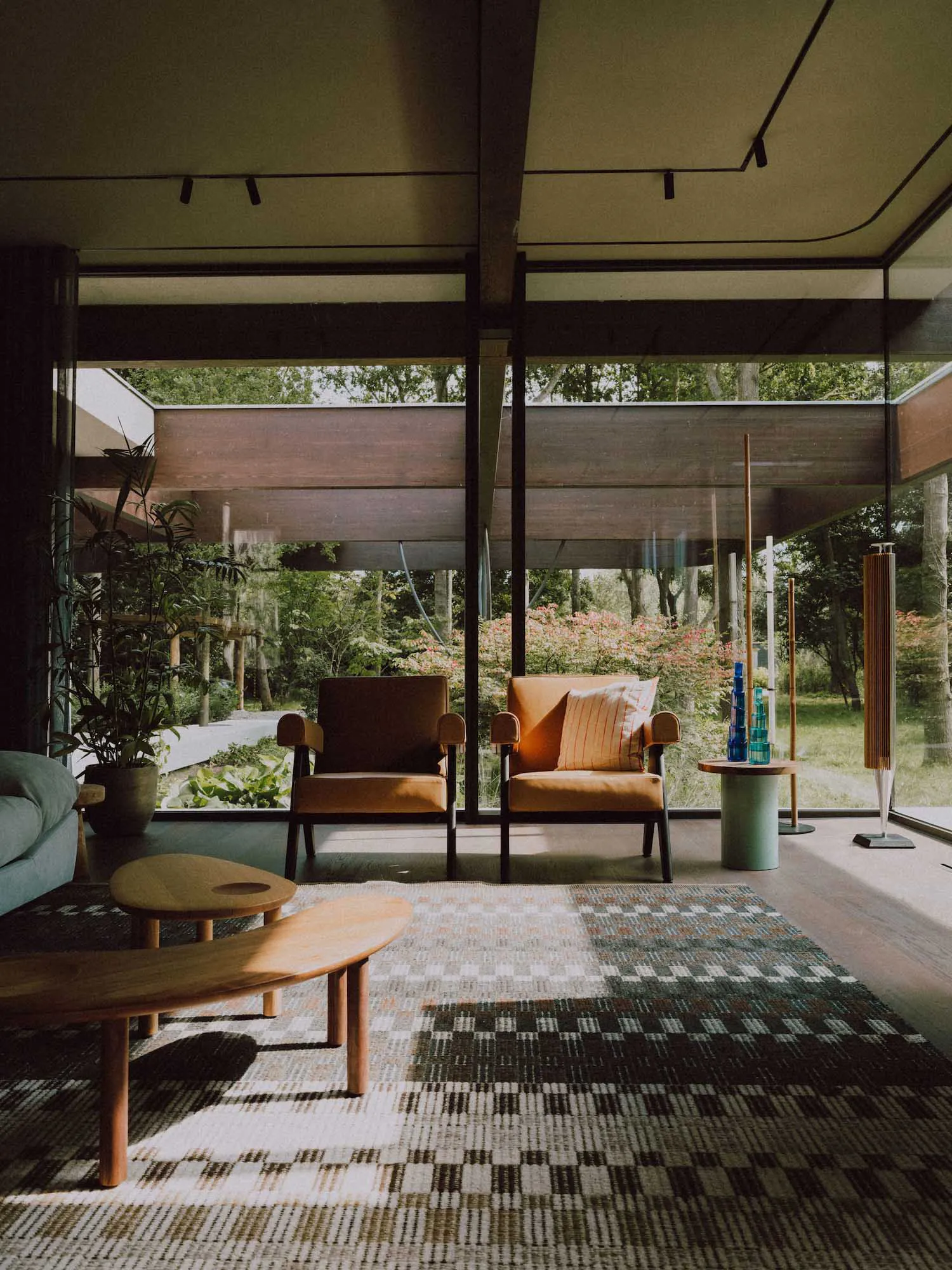 Modern living room interior featuring two orange-brown armchairs, a curved wooden coffee table, a checkered area rug, large floor-to-ceiling windows with garden views, and a potted plant.