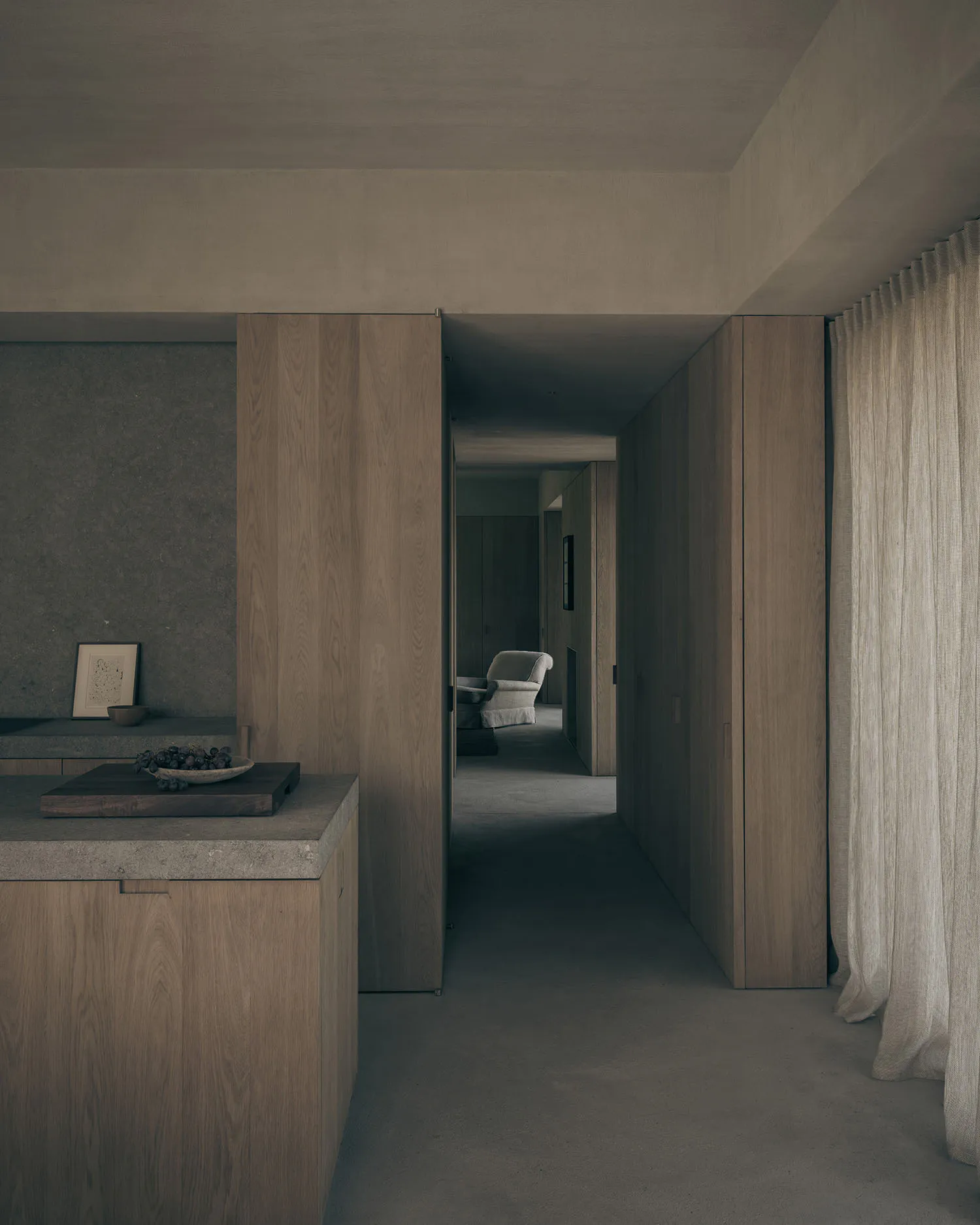 Modern minimalist hallway featuring floor-to-ceiling oak cabinetry, a concrete kitchen island with a wooden cutting board and fruit bowl in the foreground, textured concrete walls, and a view into a distant room containing a light-colored armchair.
