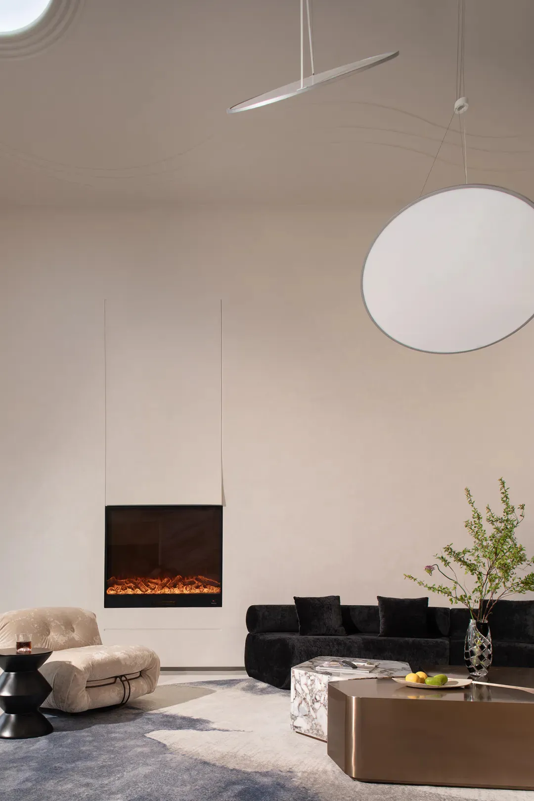 Modern minimalist living room featuring a black velvet sectional sofa, cream bouclé lounge chair, floating electric fireplace, marble and bronze coffee tables, and large circular pendant lights against curved white walls.