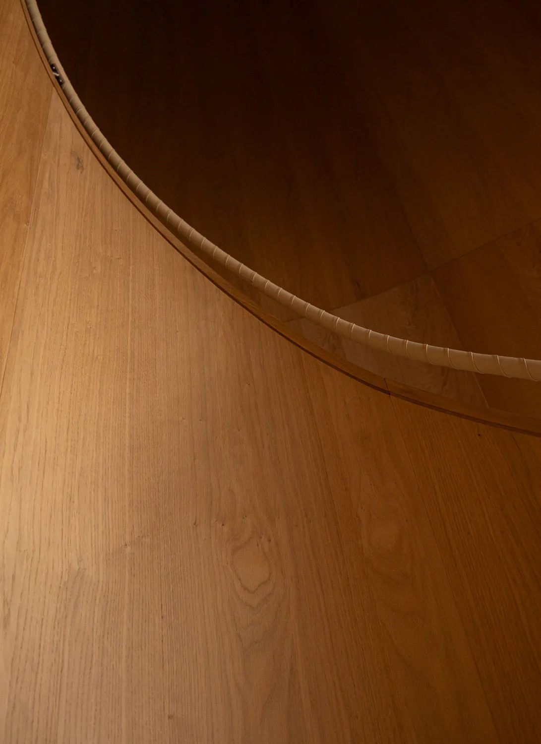 Close-up photograph of the curved edge of a thick solid oak round dining table positioned over a light oak wood plank floor, showing the smooth tabletop and ribbed side detail.