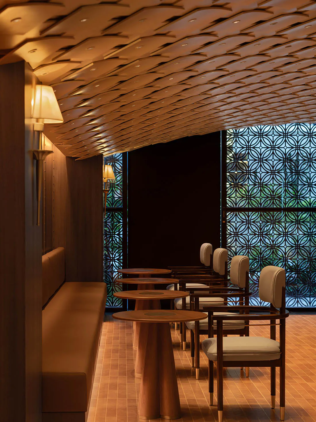 Interior view of a sophisticated Japanese-style restaurant featuring a long upholstered banquette, round wooden pedestal tables, and matching armchairs with cream upholstery, all beneath an elaborate sculptural wooden ceiling with integrated lighting and decorative lattice window screens in the background.