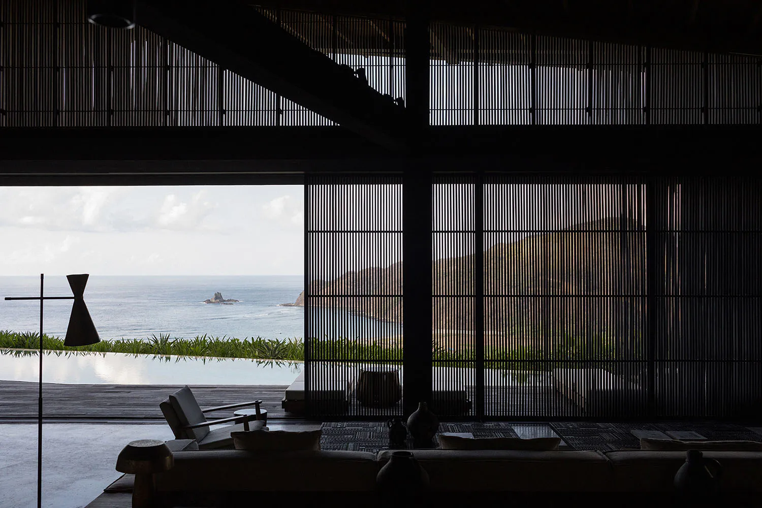 Modern open-plan living room interior with dark bamboo vertical screens, minimalist furniture, and a sweeping view of the ocean and distant rocky island through large openings.