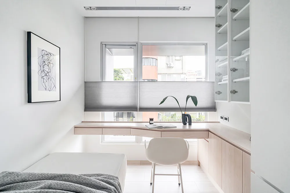 Modern minimalist home office featuring an L-shaped light wood desk, white ergonomic chair, built-in storage cabinets, large window with blinds, and a single bed in a bright, clean room.