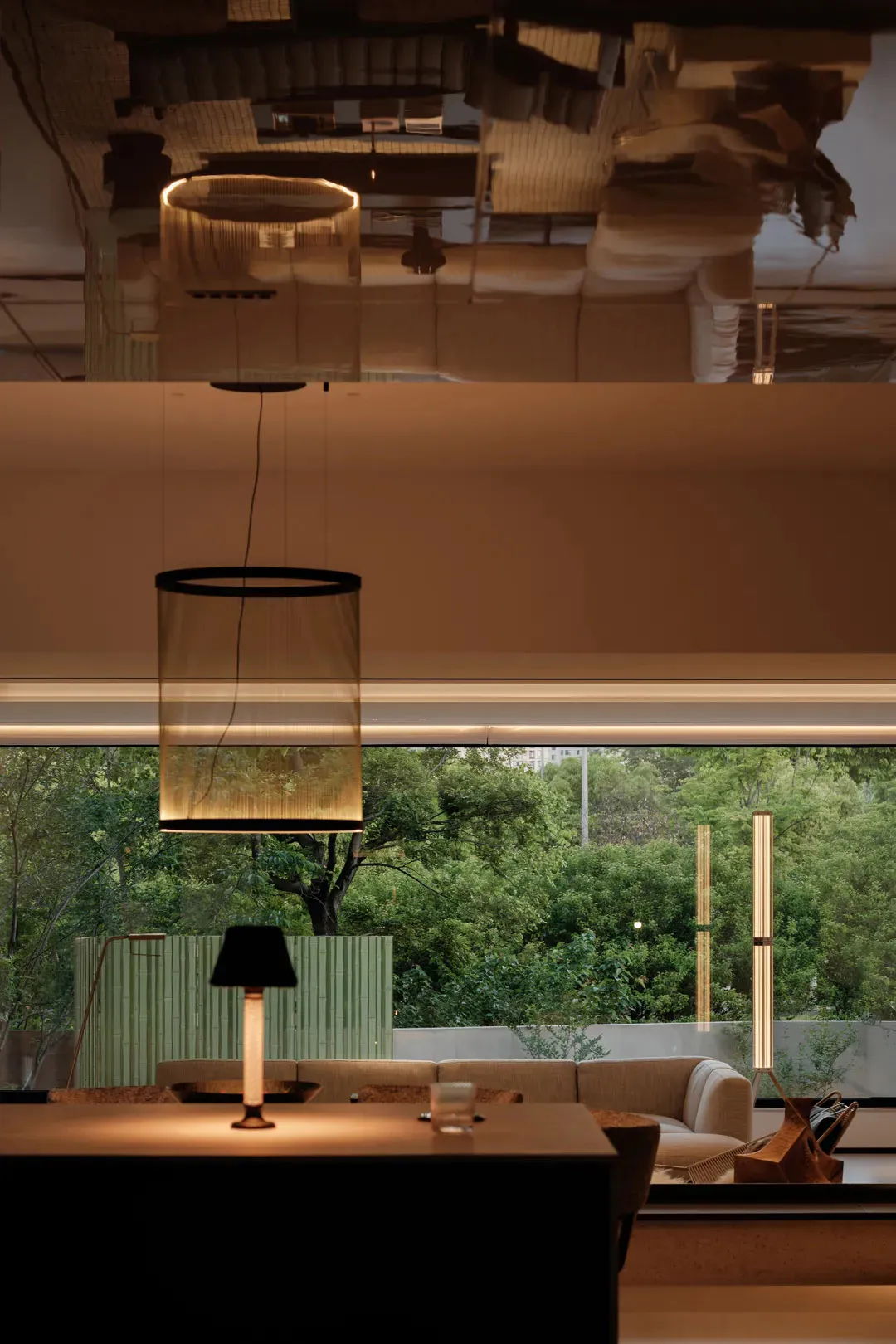 Modern living room interior featuring a large cylindrical sheer fabric pendant light, neutral sofa, table lamp, and expansive windows overlooking dense green trees.