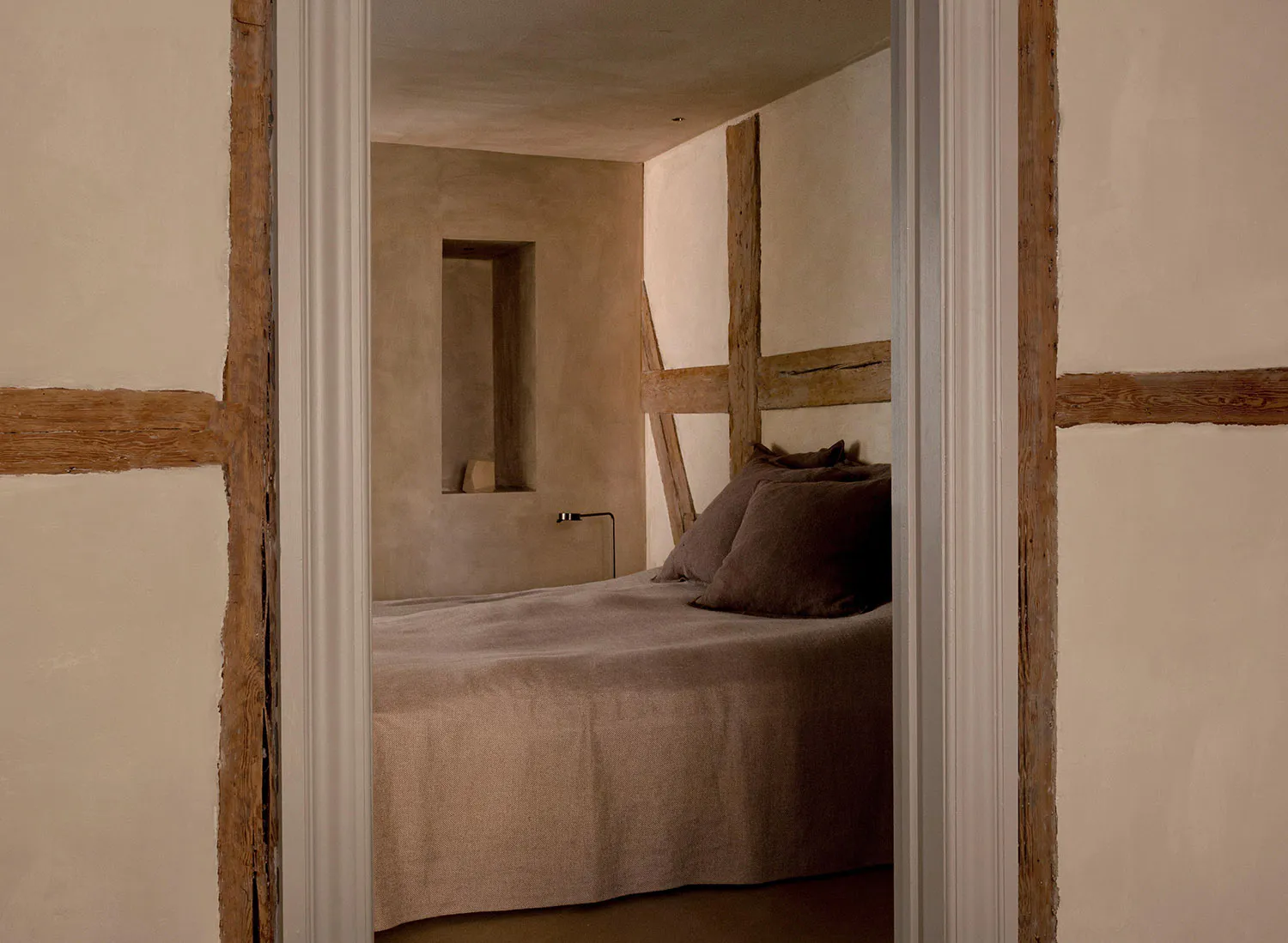View through a doorway into a minimalist wabi-sabi style bedroom with exposed wooden beams, textured plaster walls, and a bed dressed in earthy brown linen bedding.