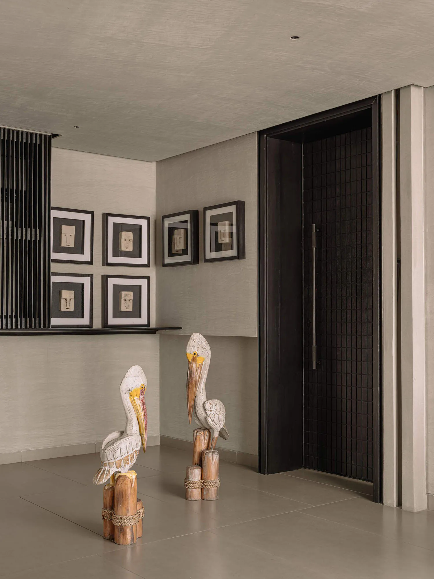 Modern minimalist entryway featuring two large vintage wooden pelican sculptures, framed tribal masks on the wall, and a dark textured front door.