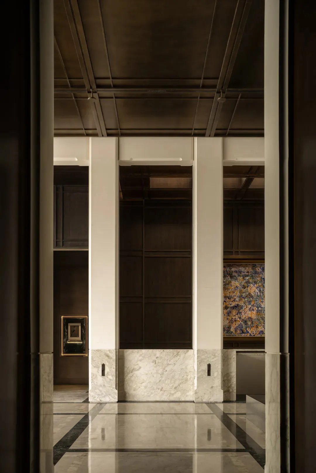View through a doorway into an elegant building lobby with white marble wainscoting, dark wood paneling, polished geometric floor tiles, and a colorful abstract painting on the right wall.