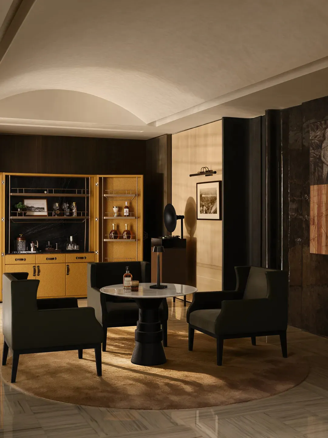 Elegant modern interior lounge with four dark green armchairs around a round white marble table on a black pedestal, set on a circular beige rug. In the background is a luxurious brass and black marble bar cabinet with glassware and spirits, dark wood paneling, and dramatic wall sconce lighting.