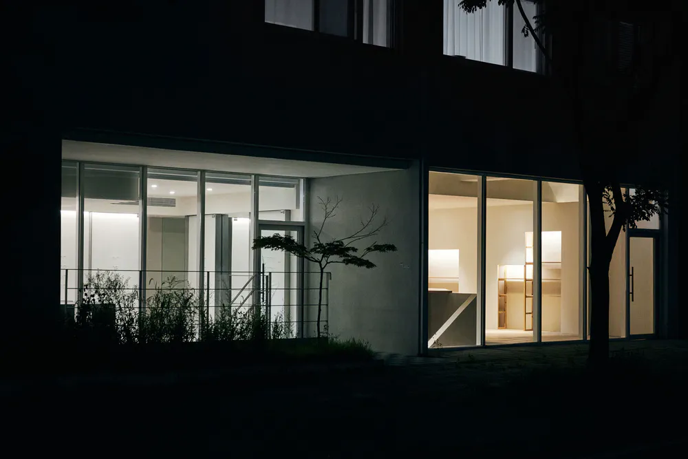 現代極簡混凝土住宅夜景,大面積玻璃窗透出溫暖室內燈光,周圍環繞樹木與植栽。