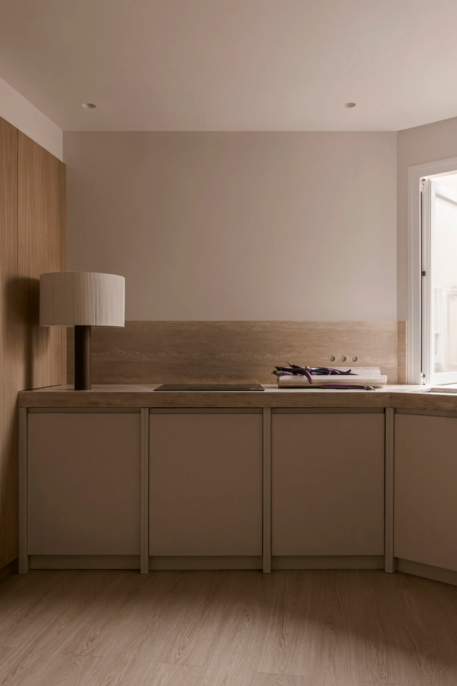 Modern minimalist kitchen with light wood cabinetry, stone countertop, wooden backsplash, table lamp, and fresh eggplants on a cutting board near a window.