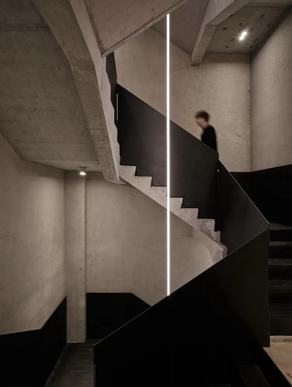 Modern brutalist concrete staircase with black metal railings, dramatic vertical linear light, and a person walking upstairs in a minimalist interior.
