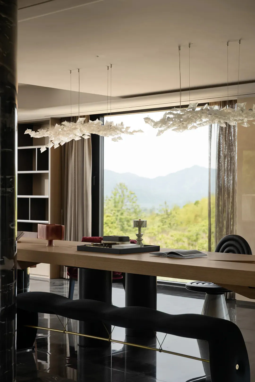 Modern dining room interior featuring a long oak table, black sculptural chairs, a black velvet bench with brass detail, and two large artistic chandeliers resembling flocks of white butterflies, with a panoramic view of green mountains through floor-to-ceiling windows.