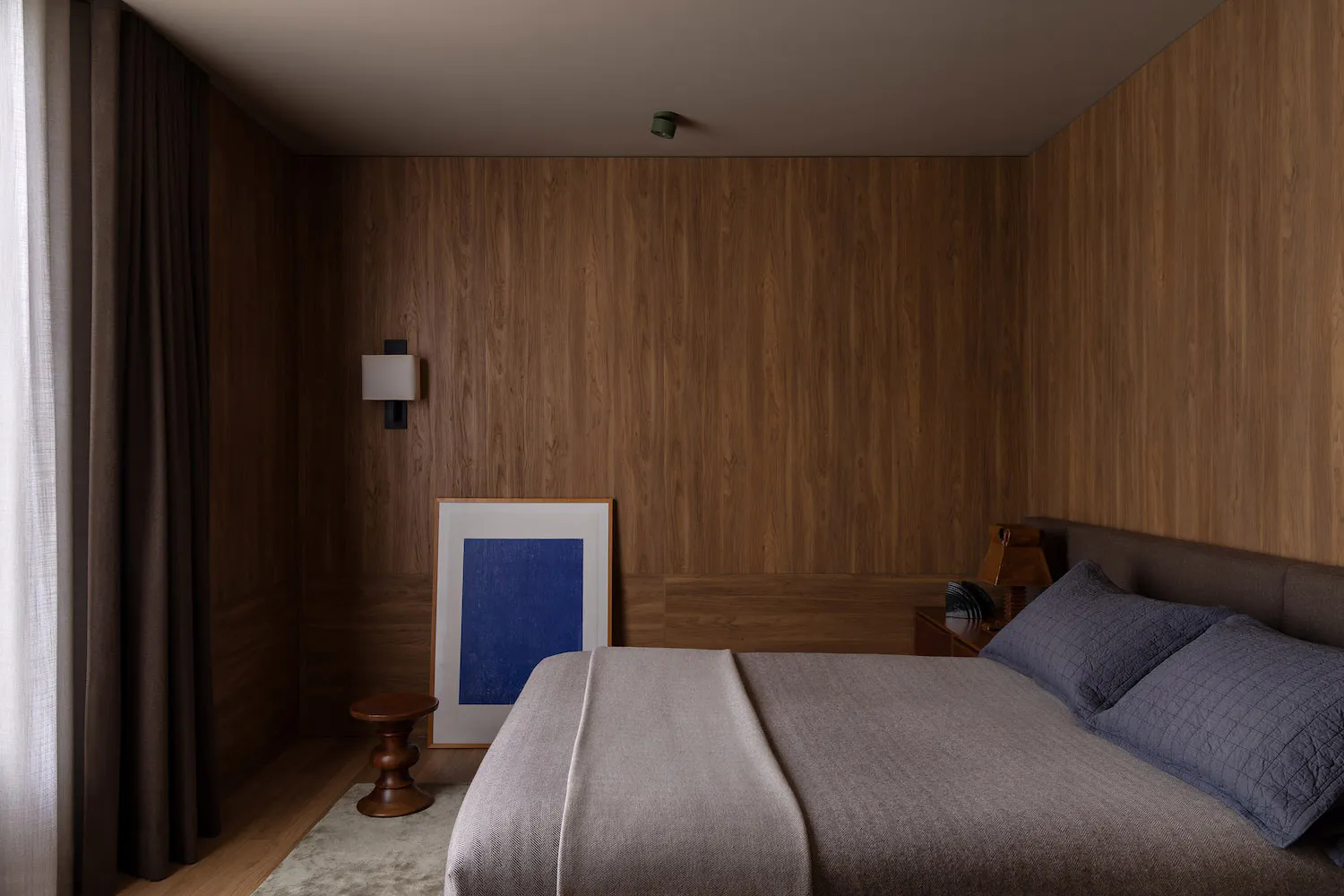 Modern minimalist bedroom with walnut wood walls, a large bed with grey bedding and blue pillows, a wooden side table, blue abstract framed art, and layered curtains.