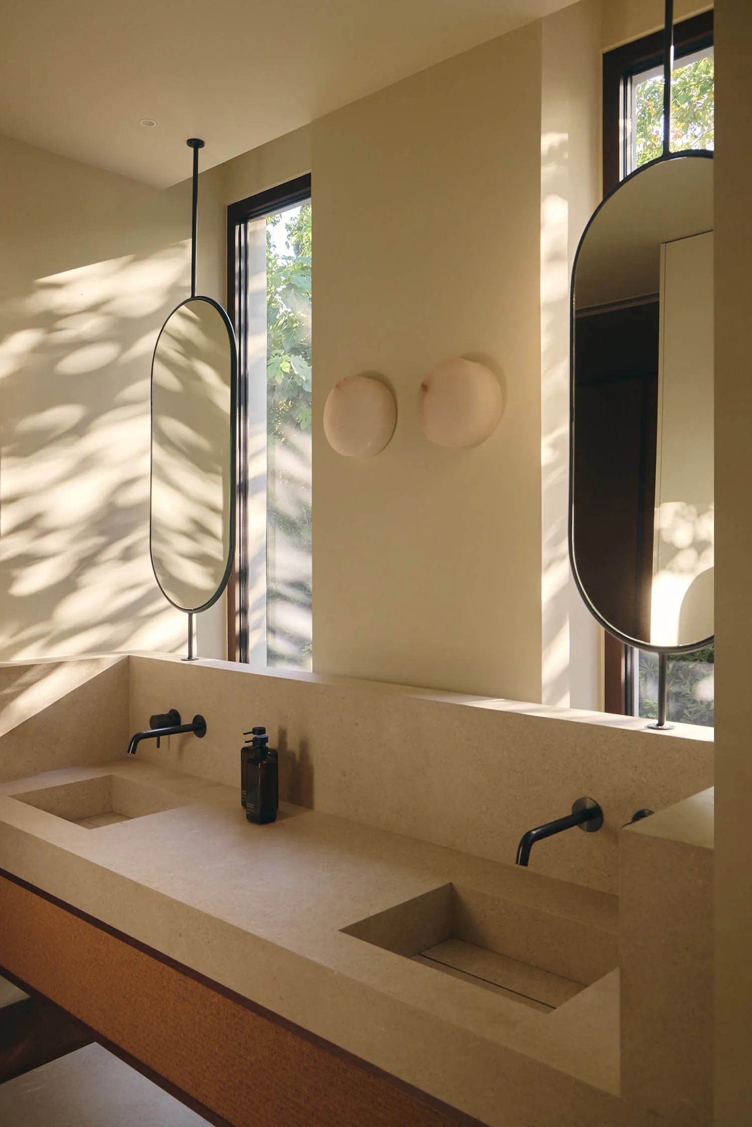 Modern minimalist bathroom with a long terrazzo countertop featuring two integrated rectangular sinks, black wall-mounted faucets, two oval mirrors suspended from the ceiling, and two spherical wall sconces, illuminated by soft natural light from large windows showing green foliage outside.