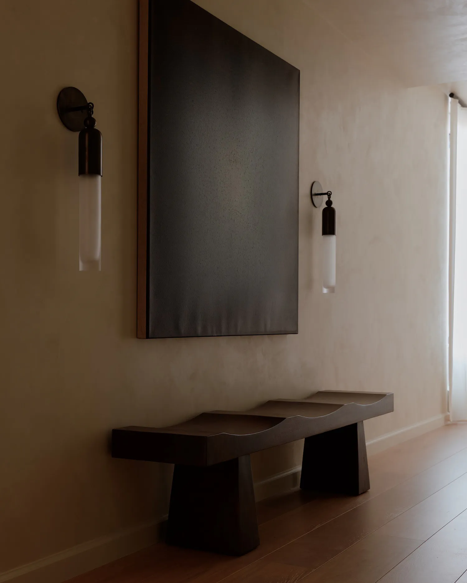 A modern minimalist entryway featuring a dark wavy walnut console table beneath a large textured dark abstract painting, flanked by two matching modern wall sconces with cylindrical glass shades, set against warm beige plaster walls and light wood flooring.