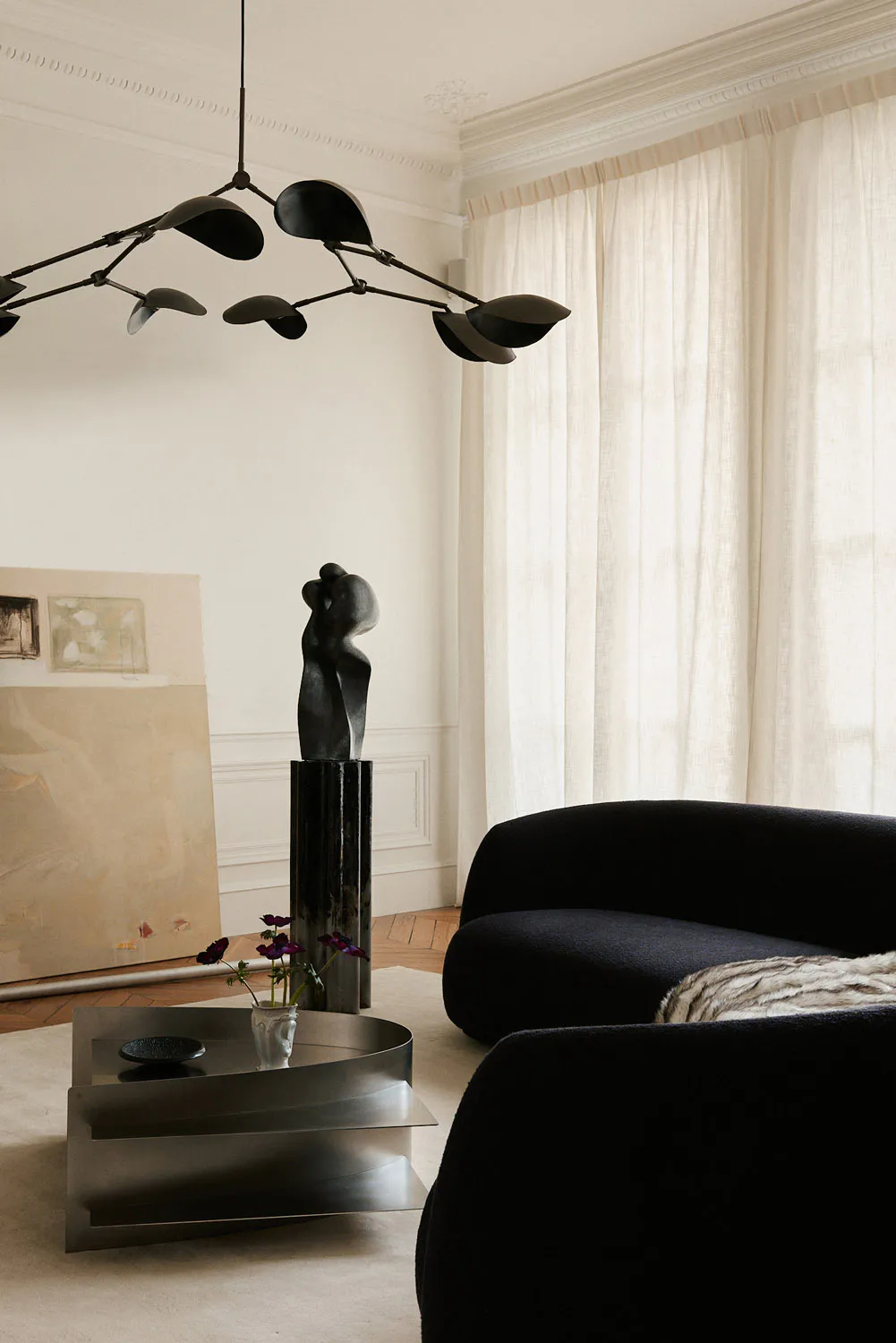 Modern living room interior featuring a large black velvet curved sofa, a multi-arm black kinetic chandelier, a dark abstract sculpture on a tall pedestal, a metallic tiered coffee table with purple flowers, and large abstract paintings leaning against a white wall with crown molding.