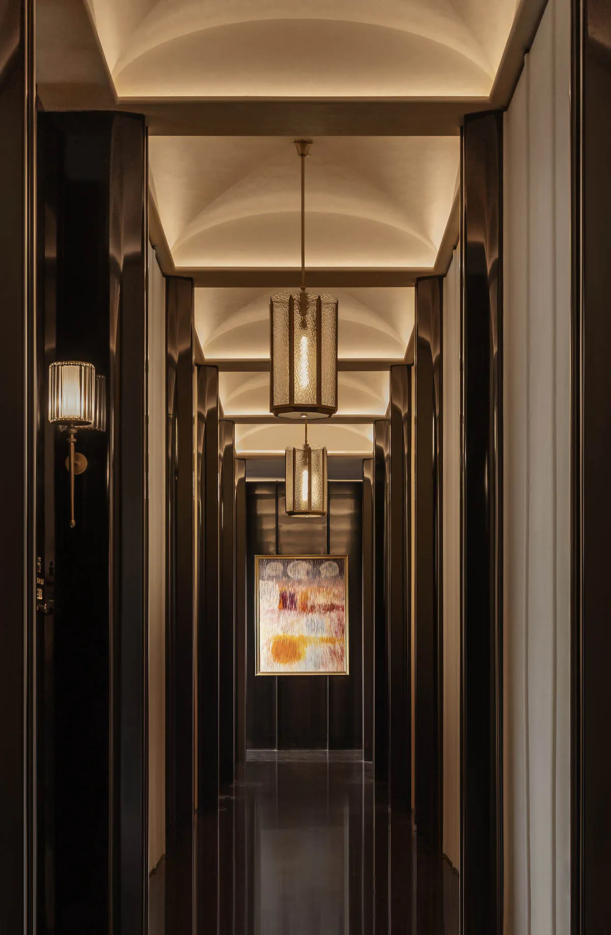Long narrow luxury hotel hallway with dark wood paneling, arched ceilings with cove lighting, textured cylindrical pendant lanterns, and a colorful abstract painting at the end.