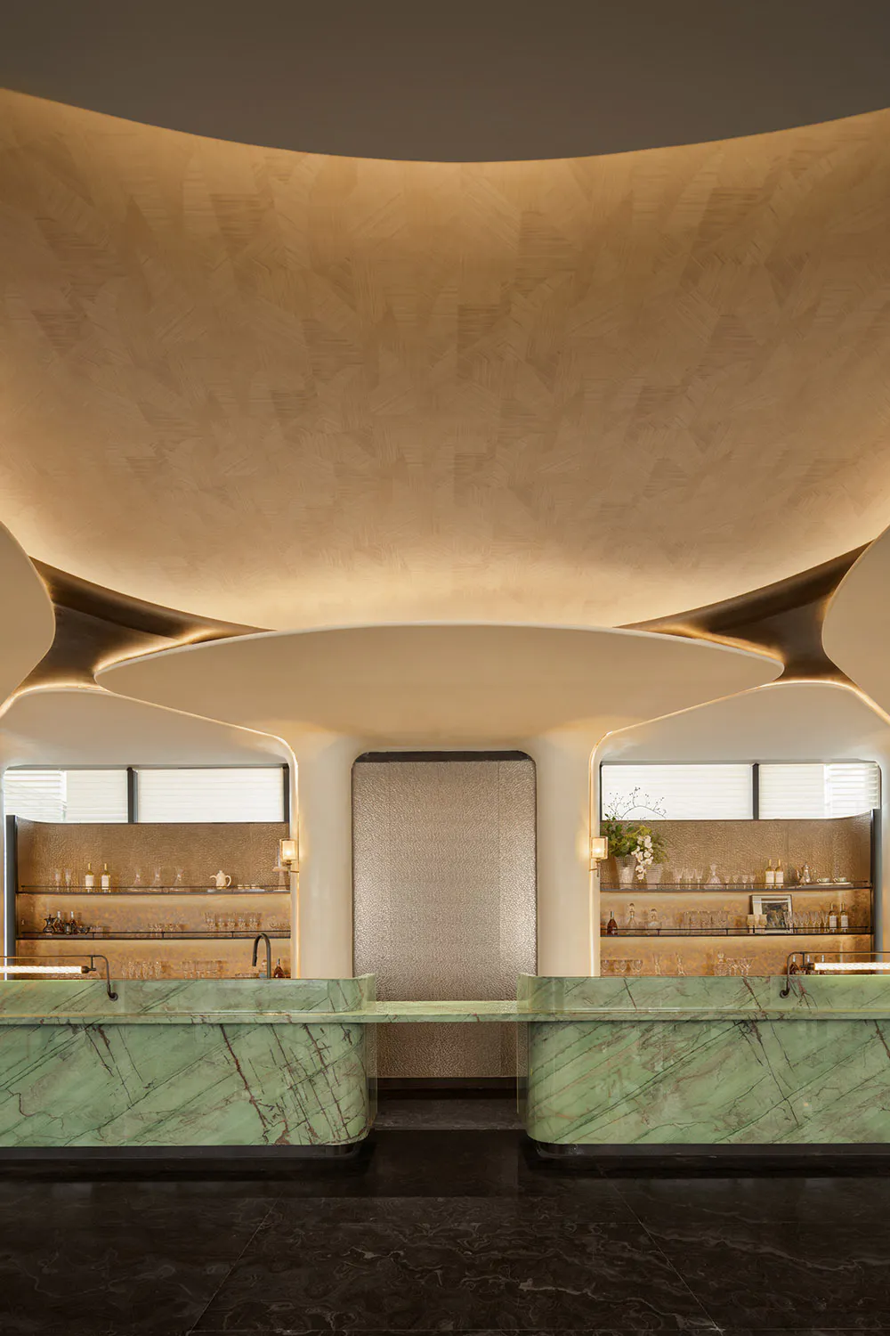 Modern luxury bar reception with green marble counters, curved organic ceiling with warm wood texture, metallic central wall panel, and backlit shelving displaying glassware.