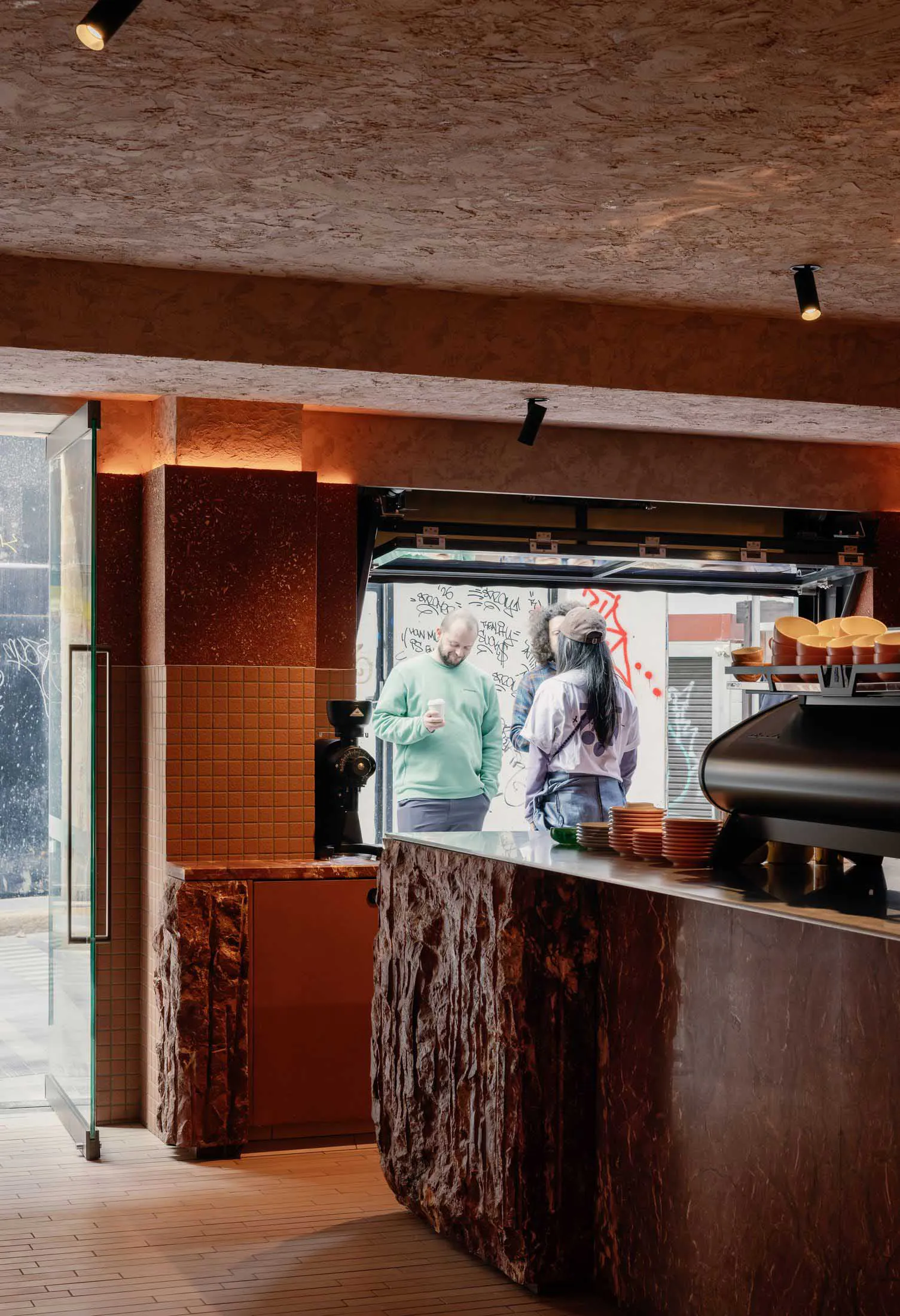Interior view of a modern coffee shop with a dramatic service counter made from rough volcanic rock and polished terrazzo, featuring orange-brown tiled walls, a coffee grinder, stacked ceramic cups, and customers interacting at the window-style service hatch under warm spotlighting.