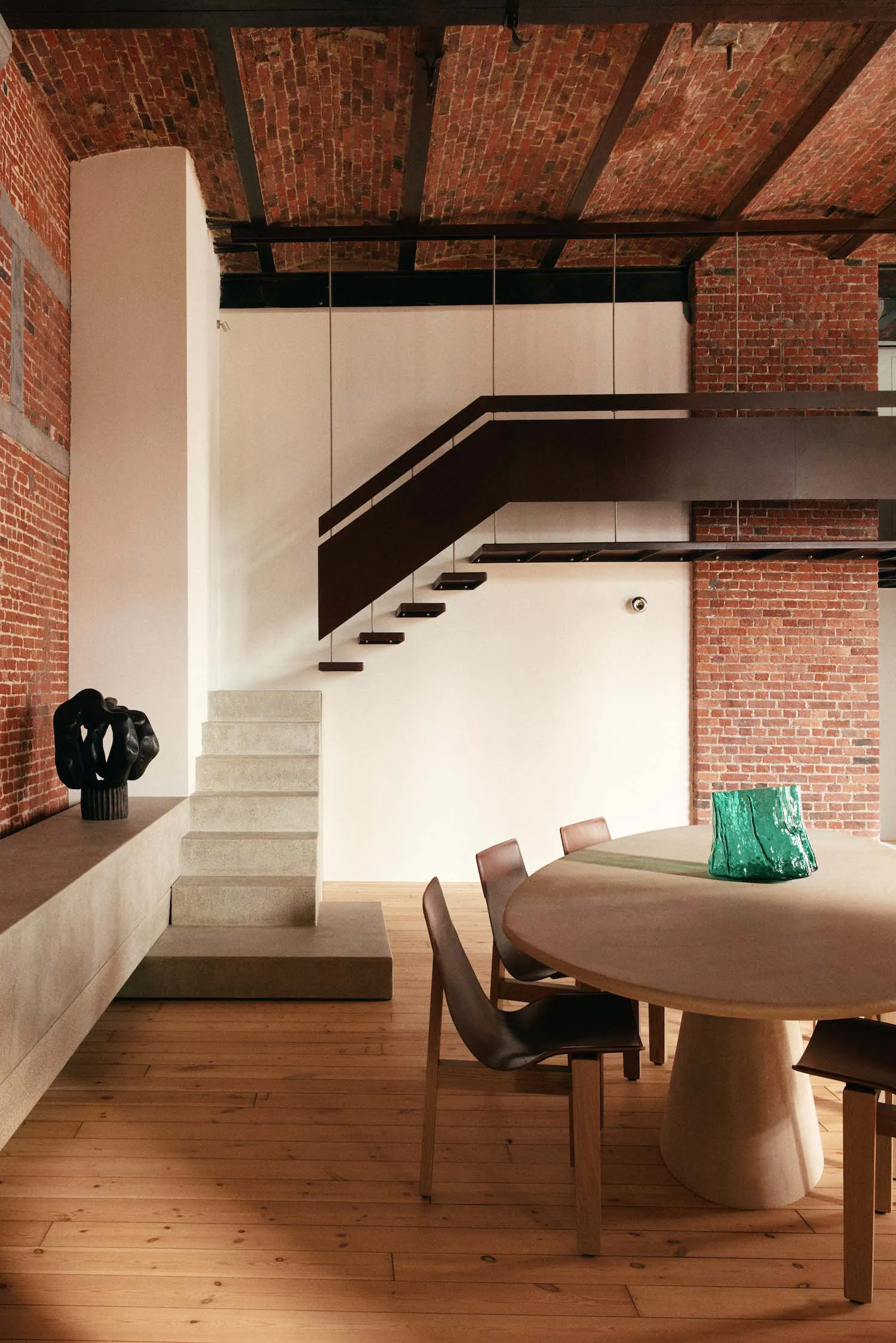 Modern industrial loft interior featuring a floating concrete staircase, exposed red brick walls, wooden floor, and a round dining table with brown leather chairs and a green glass sculpture.