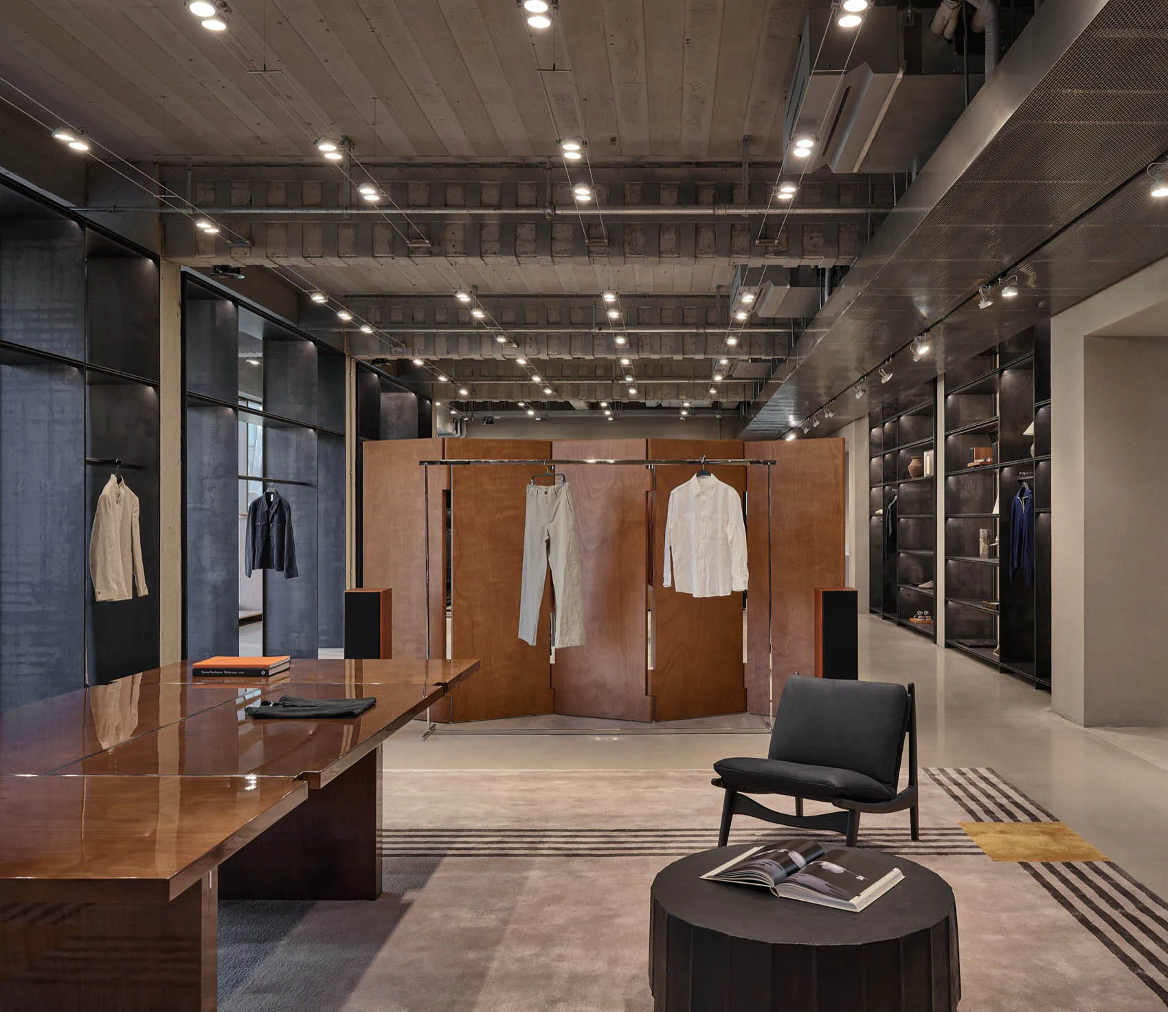 Modern industrial-style mens clothing boutique interior with polished walnut table, dark grey armchair, concrete ceiling, and clothing display racks.