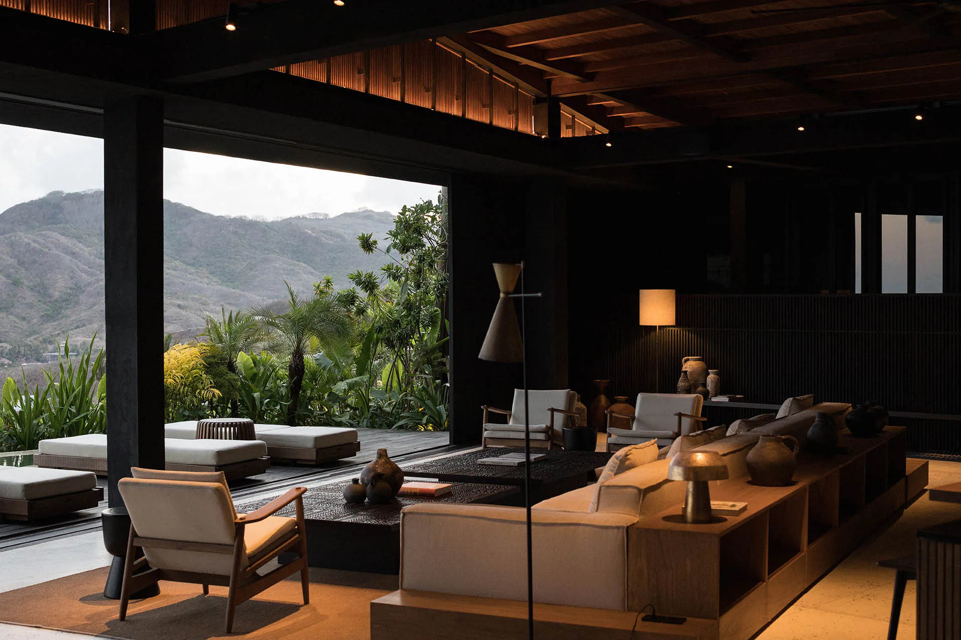 Modern open-plan living room with large glass openings overlooking green tropical mountains, featuring neutral sofas, wooden armchairs, woven coffee tables, and earthy ceramic vases.