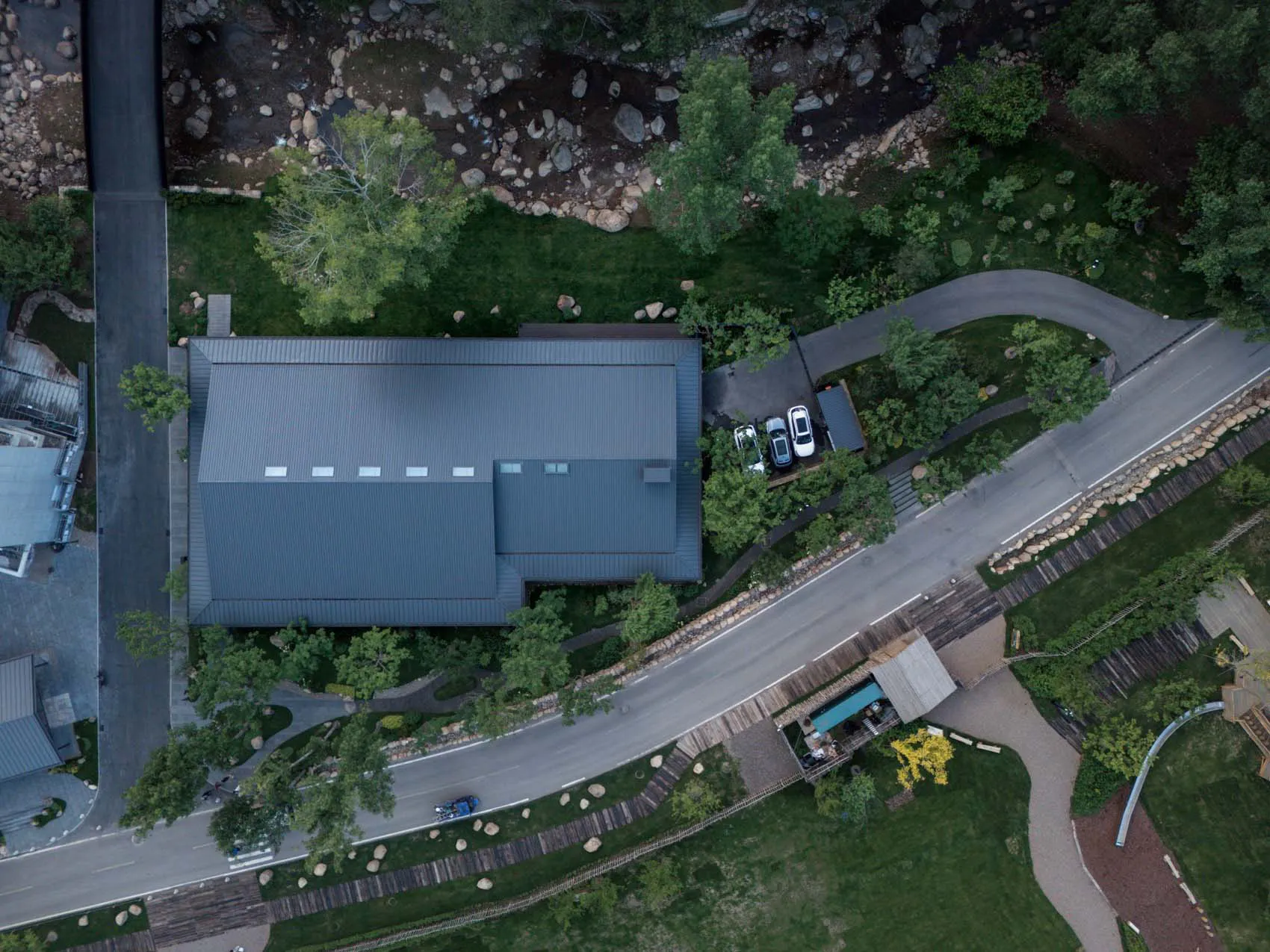 Aerial photograph of a modern rectangular house with dark standing seam metal roof, surrounded by landscaped garden, next to a rocky river and paved driveway with parked cars.