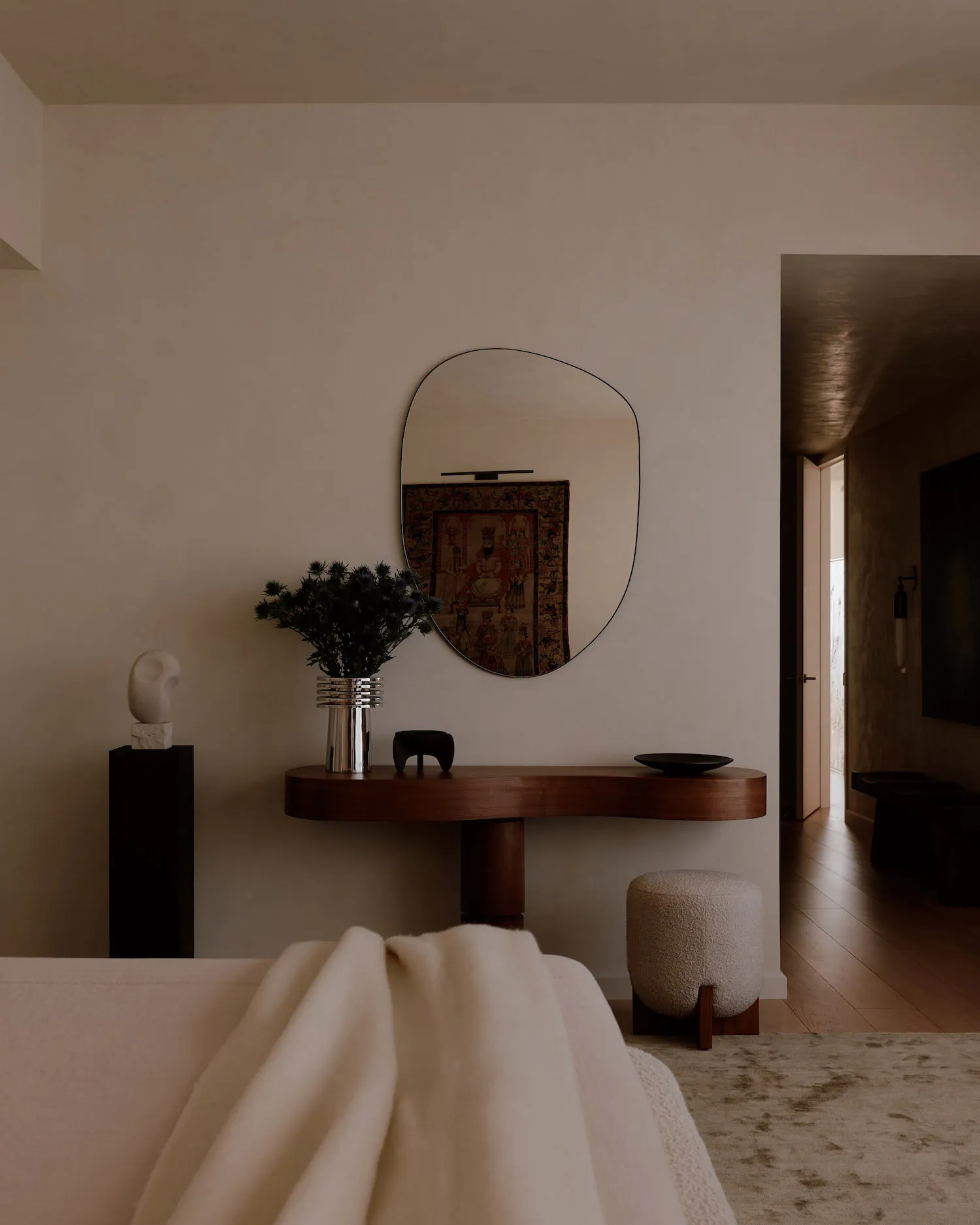 Modern minimalist entryway featuring a curved walnut console table, irregular organic mirror, metallic vase with dried flowers, white stone sculpture on a black pedestal, and a bouclé ottoman.