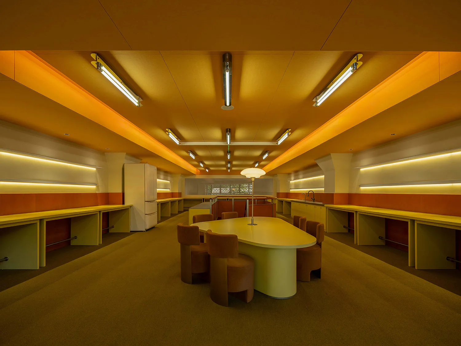 Long view of a retro mid-century modern communal kitchen with orange ceiling panels, built-in yellow counters, a large oval avocado-green table surrounded by brown upholstered chairs, and fluorescent tube lighting.