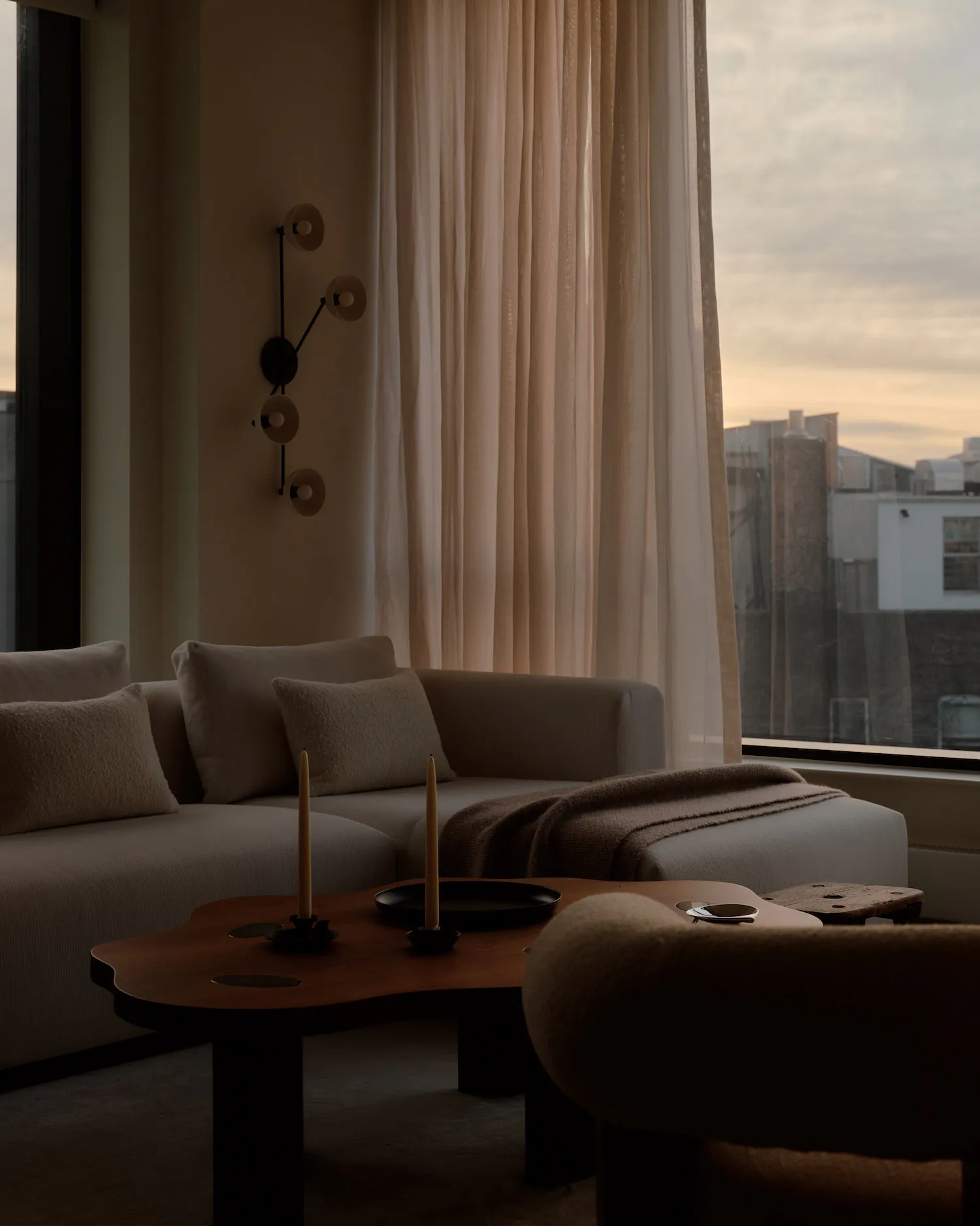 A contemporary living room featuring a light beige boucle sectional sofa with matching pillows, a dark wood irregularly shaped coffee table holding two tall candles, a modern black wall sconce with multiple circular lights, and a large window revealing a city skyline at sunset with sheer curtains.