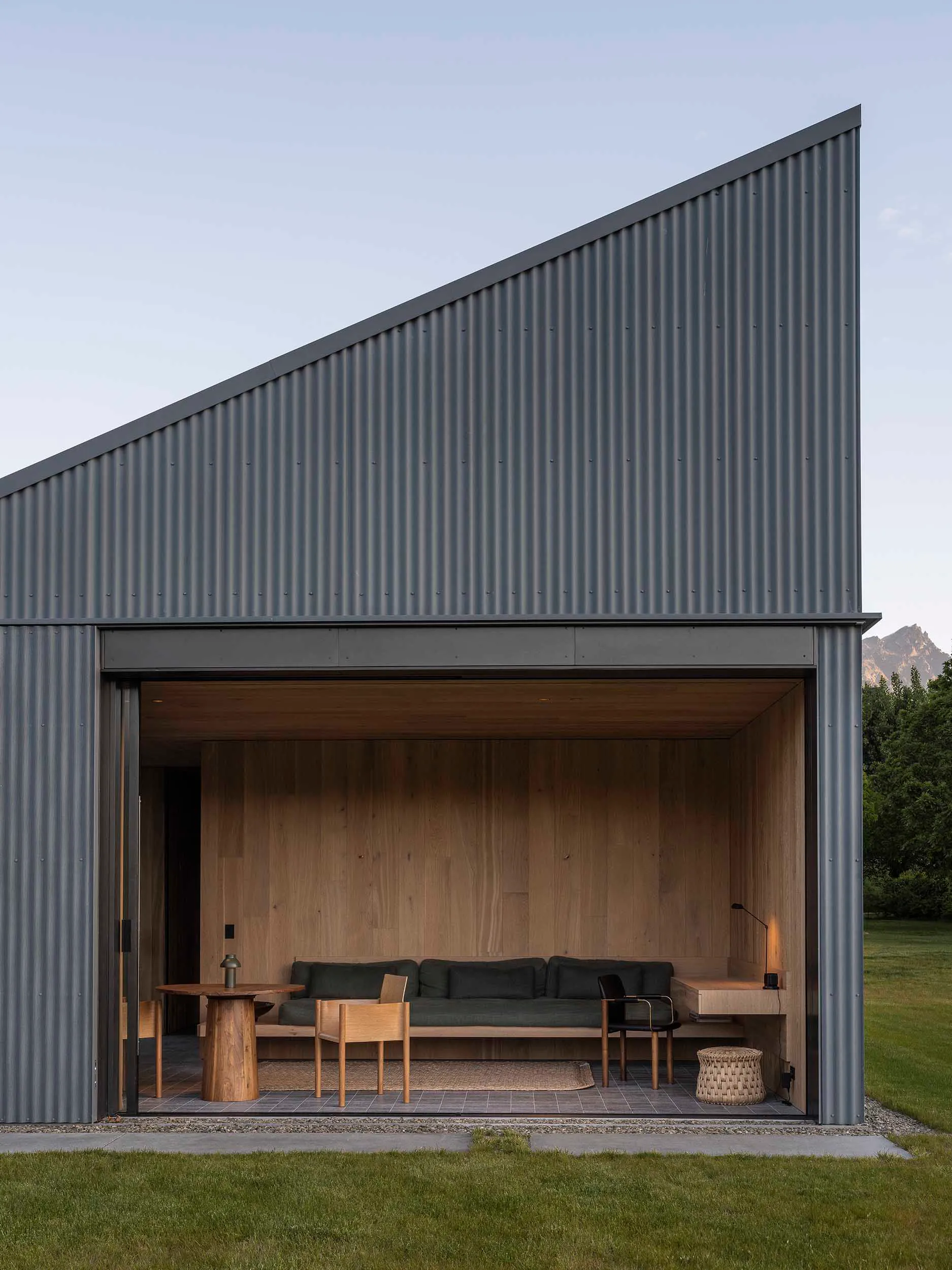 Modern cabin with dark corrugated corten steel exterior and open indoor-outdoor living area furnished with a green sofa, wooden chairs, and a round wooden table, set against a mountain backdrop.