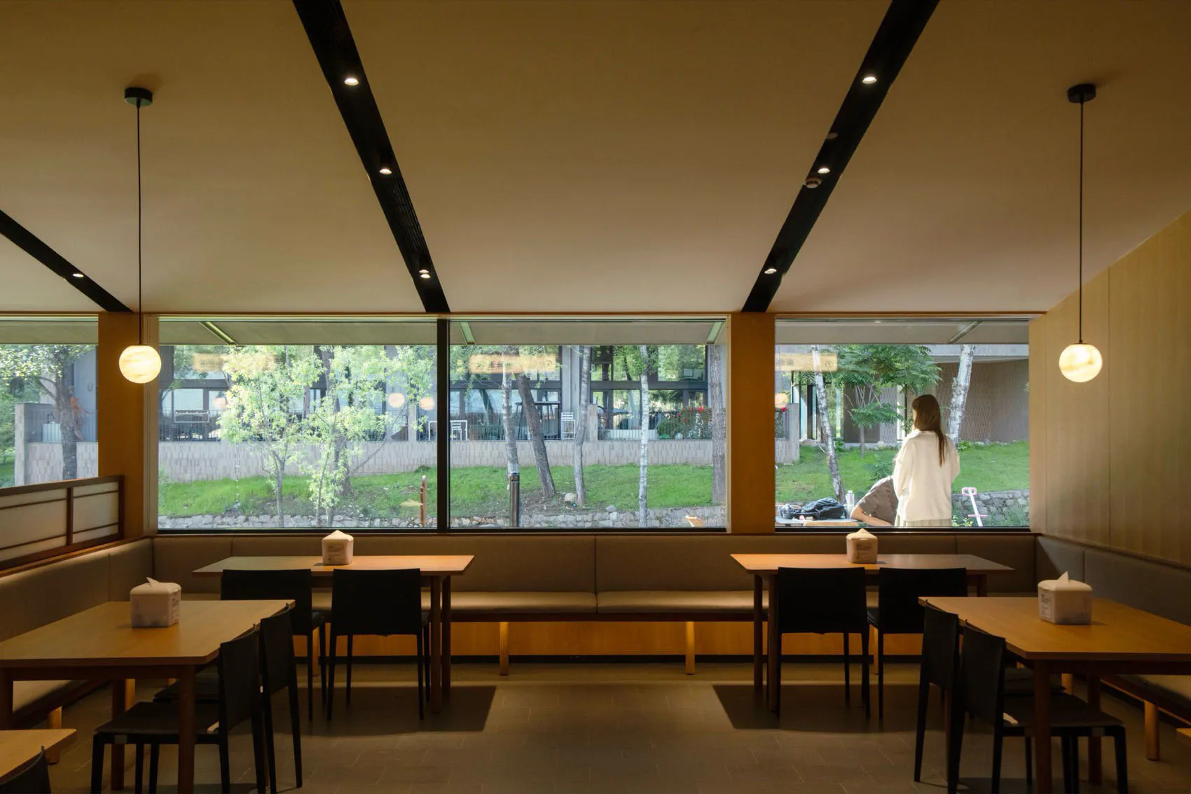 Modern minimalist Japanese restaurant interior featuring light wood tables, black chairs, built-in upholstered banquettes, large windows overlooking a green garden, and spherical pendant lights.