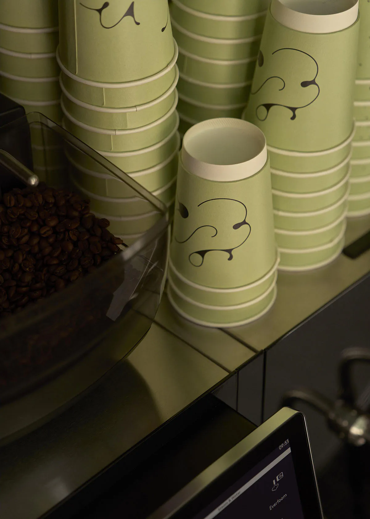 Close-up of sage green paper coffee cups with black abstract face designs stacked on a counter next to a bin of coffee beans in a cafe setting.