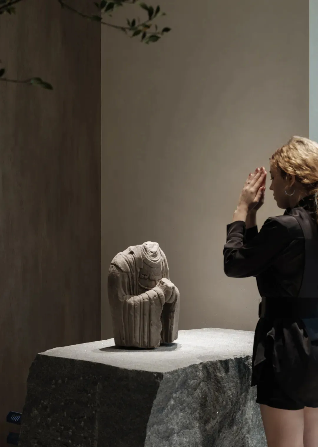 A woman with blonde hair in a black outfit stands with hands pressed together in prayer, facing a weathered headless stone Buddha statue displayed on a large granite pedestal in a minimalist interior space with neutral walls and soft lighting.