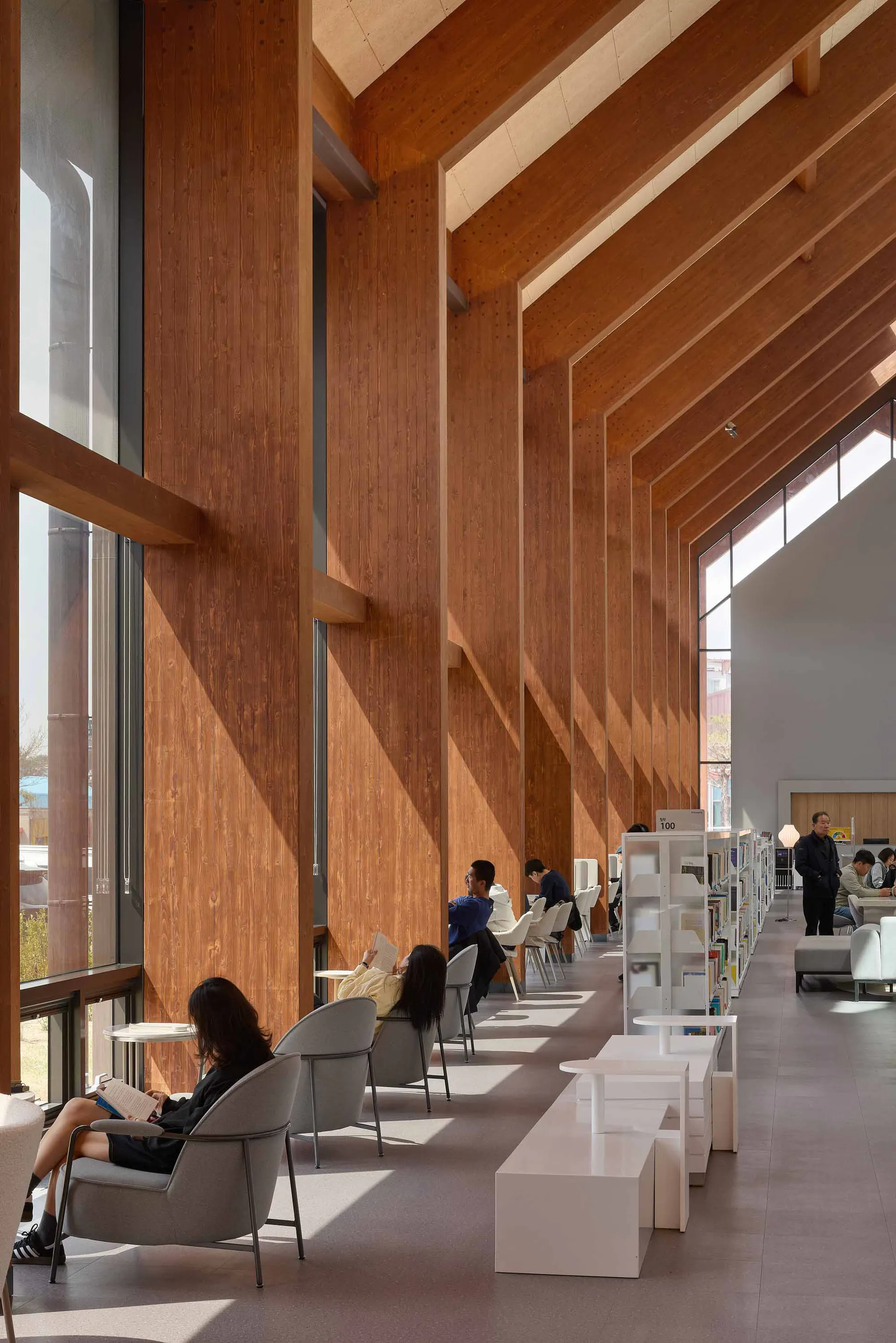 Modern library interior with tall wooden panel walls, grey reading chairs, white modular tables, and people reading in natural sunlight.