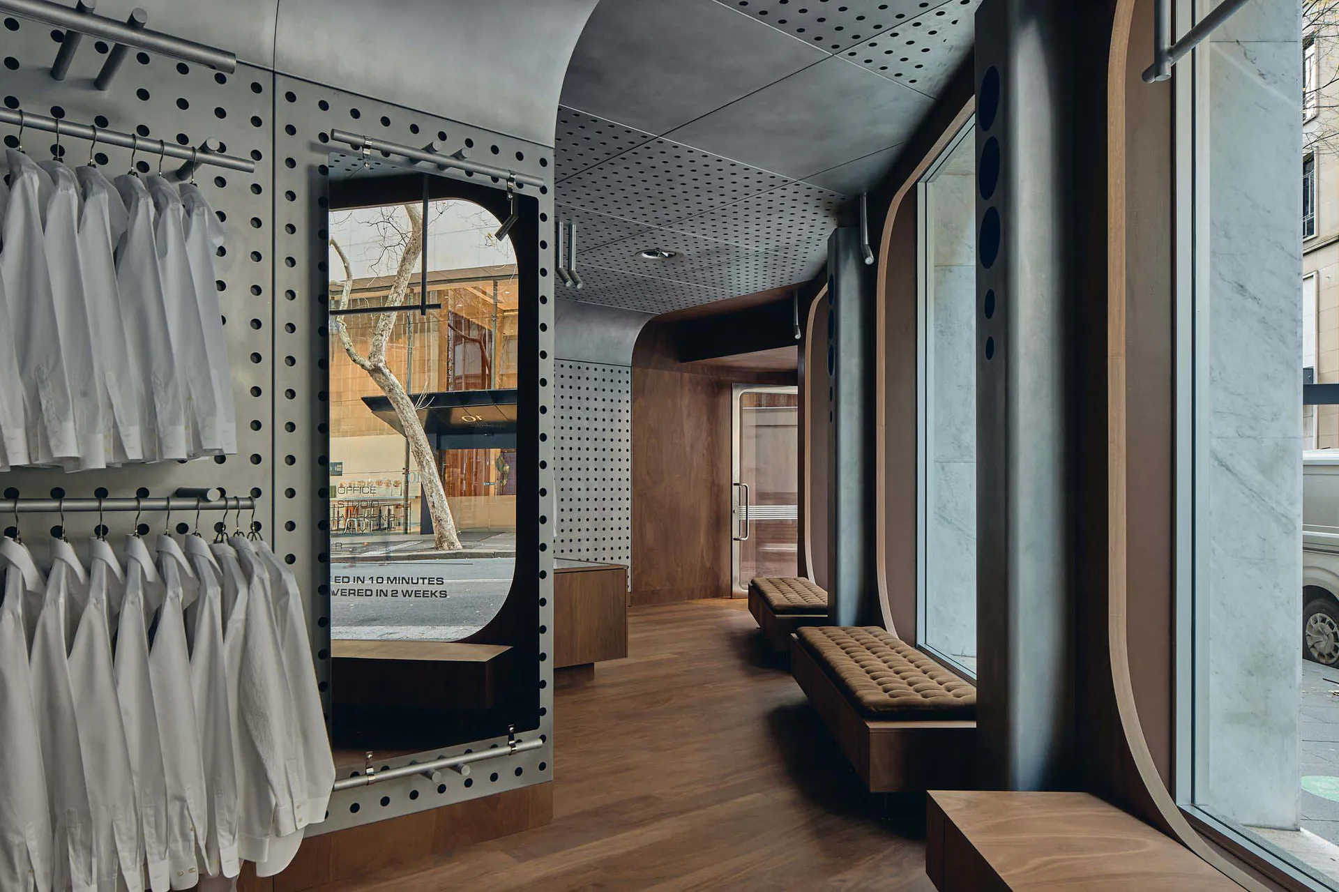 Modern boutique store interior with curved walnut wood walls, built-in upholstered benches, perforated steel clothing racks holding white shirts, and large arched windows overlooking the street.