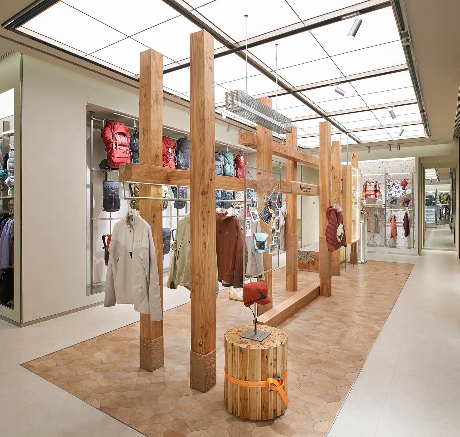 現代零售空間內部,大型回收木材柱梁結構作為服裝陳列系統,展示多款戶外機能外套與背包,搭配石紋地板與天窗天花板。