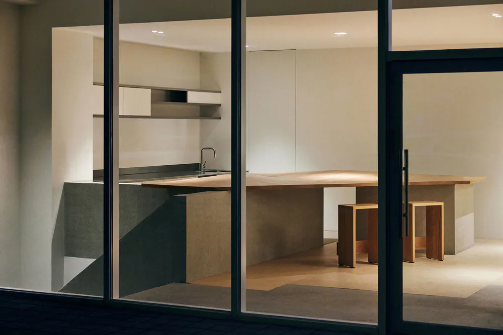 Modern minimalist kitchen viewed through glass doors at night, featuring a large live-edge oak kitchen island with concrete base, floating shelves, and soft recessed lighting.