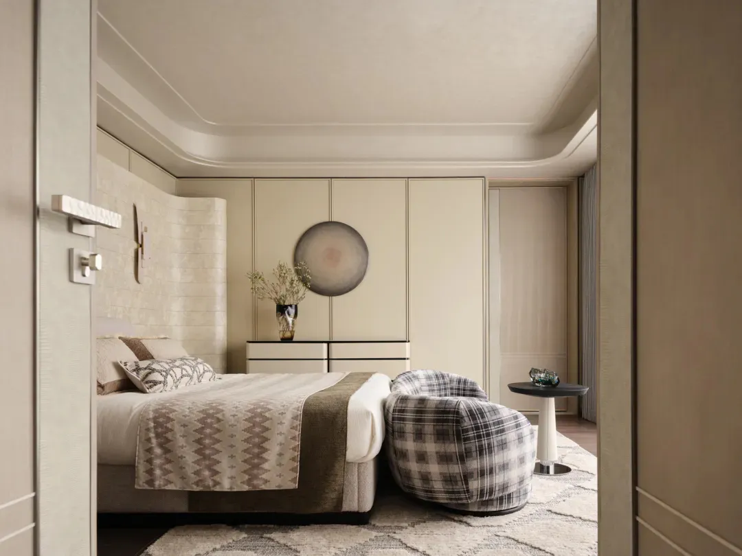 View into a modern luxury bedroom with a neutral color palette, curved headboard, plaid lounge chair, and built-in cabinetry.