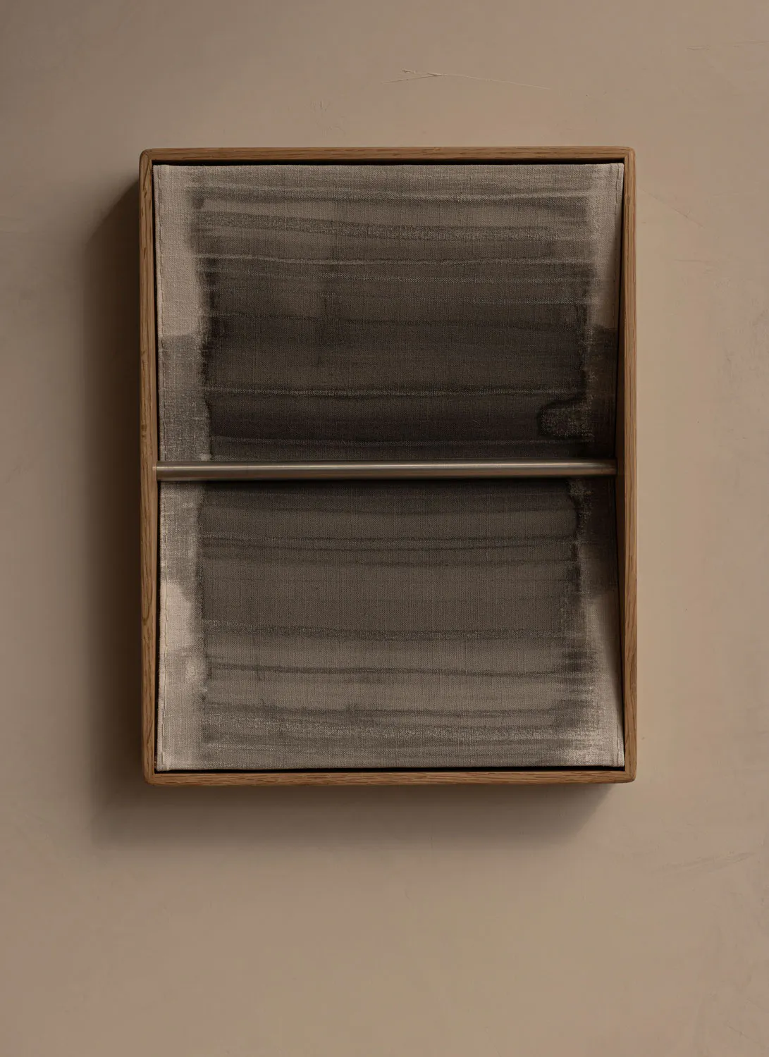 A rectangular wall-mounted artwork with a light oak wooden frame containing two panels of sheer grey fabric with horizontal brushstroke-like patterns and a horizontal metal bar dividing the upper and lower sections, hanging on a textured beige wall.
