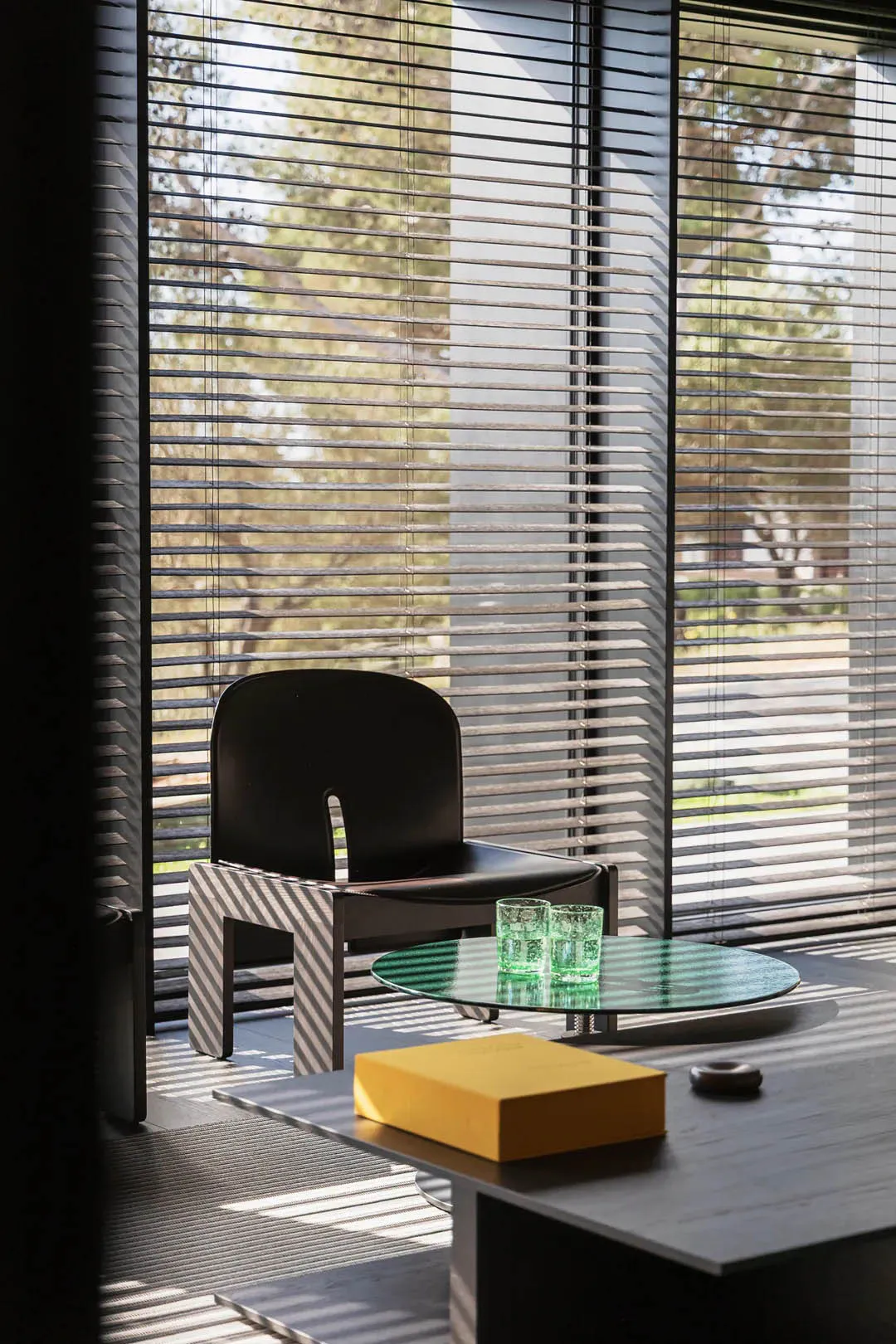 Modern black leather armchair with a distinctive U-shaped cutout back and black-and-white diagonally striped wooden base, positioned next to a round green-tinted glass coffee table holding two textured green glasses, with a yellow box and small wooden object on a dark table in the foreground, all set against large windows with horizontal blinds casting striped shadows.