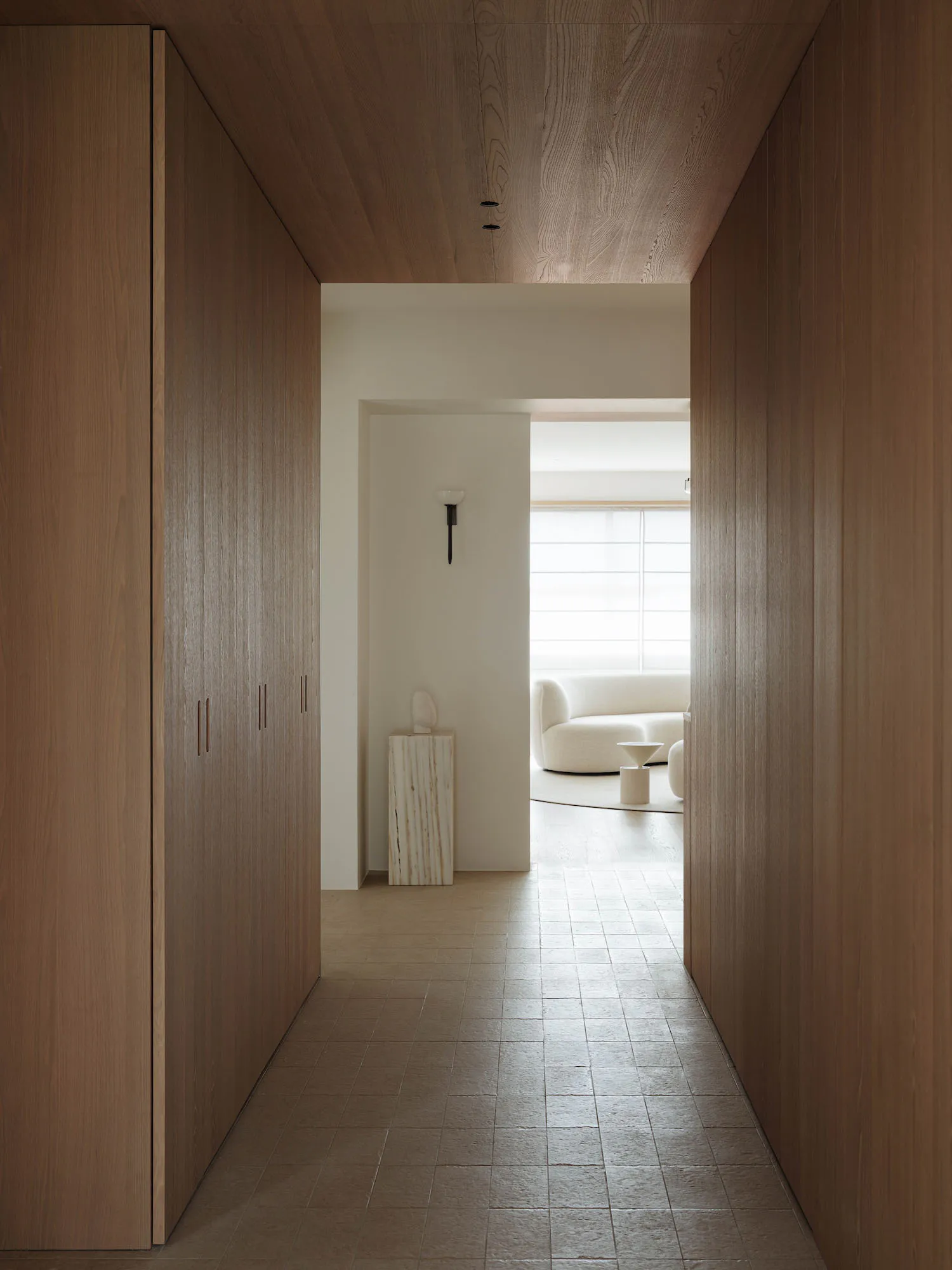 Long narrow hallway with floor-to-ceiling oak wood cabinetry and matching wood ceiling, leading to a bright living room with a curved white sofa and marble pedestal.